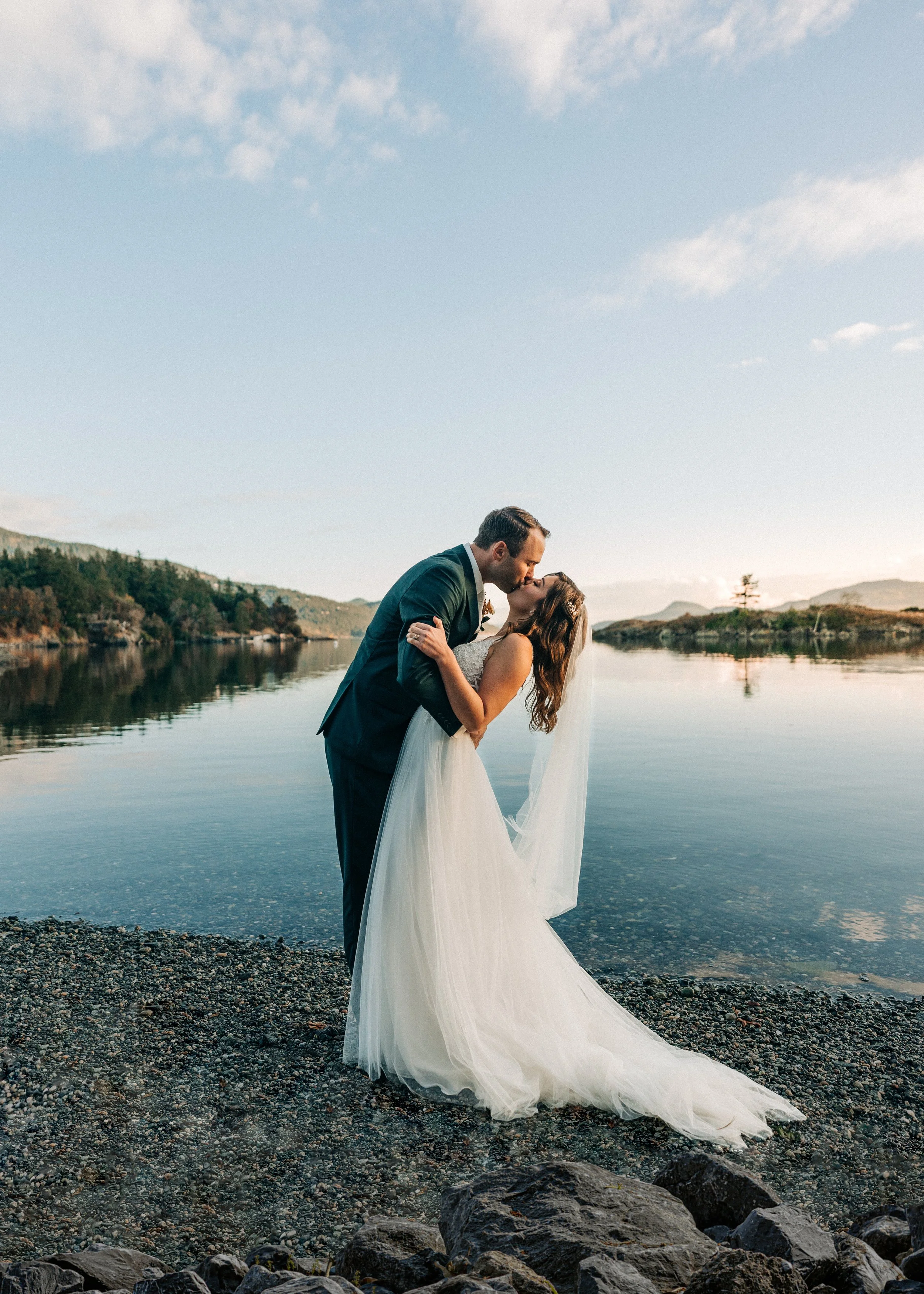 A bride and groom kissing on a rocky shore by a calm lake with mountains and trees in the background.