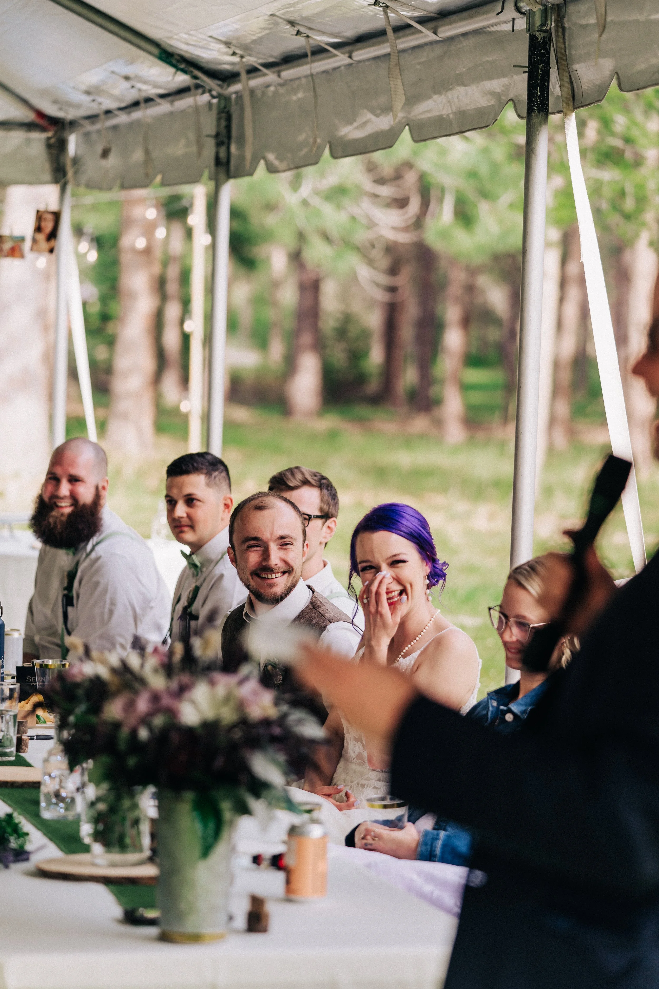 A group of people sitting at a table under a tent, smiling and laughing, with a woman in purple hair at the center of the image.
