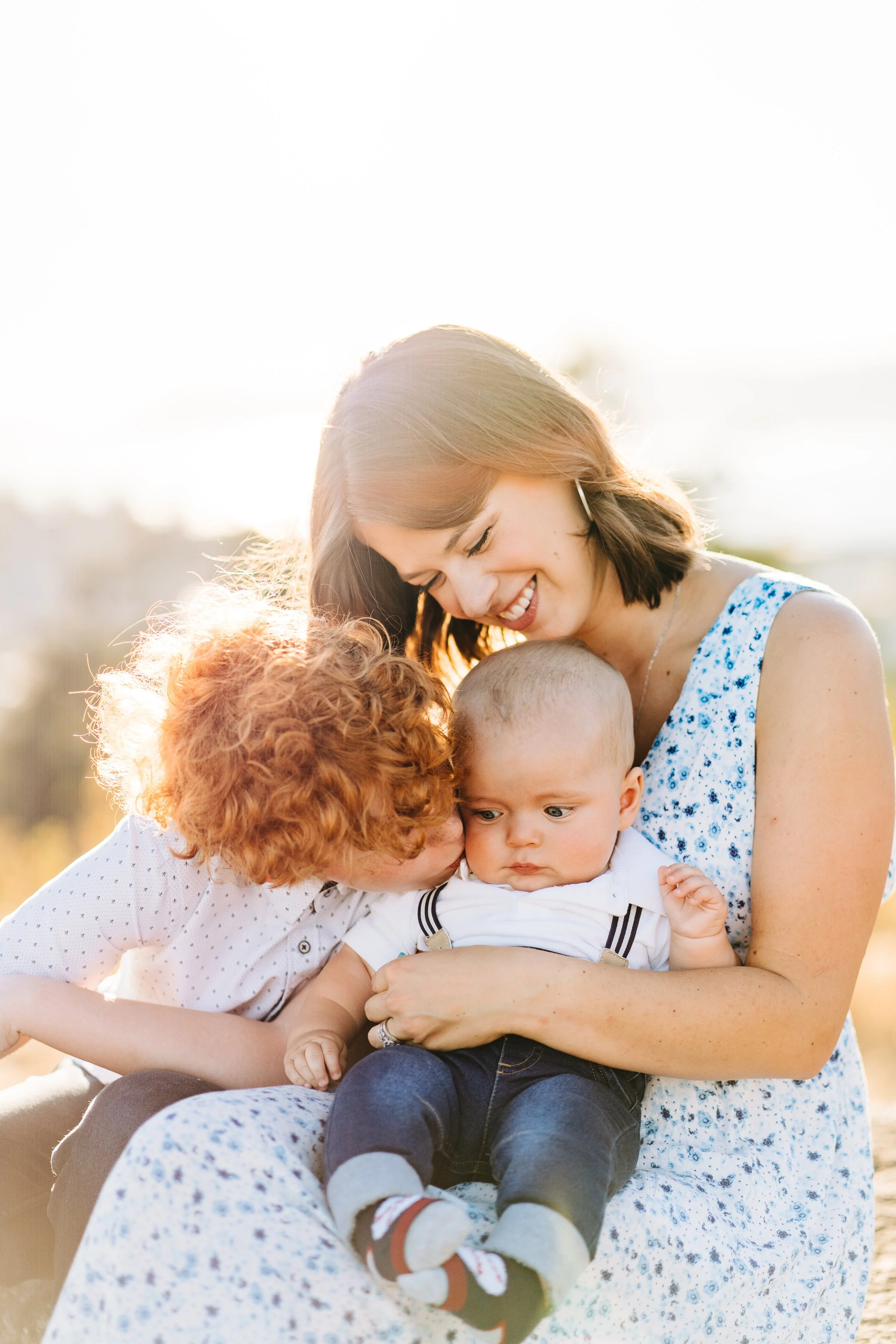 A woman with short brown hair smiling as she holds a baby boy on her lap, a girl with curly red hair kissing the baby's cheek in an outdoor setting with sunlight | Chelan Family Photographer