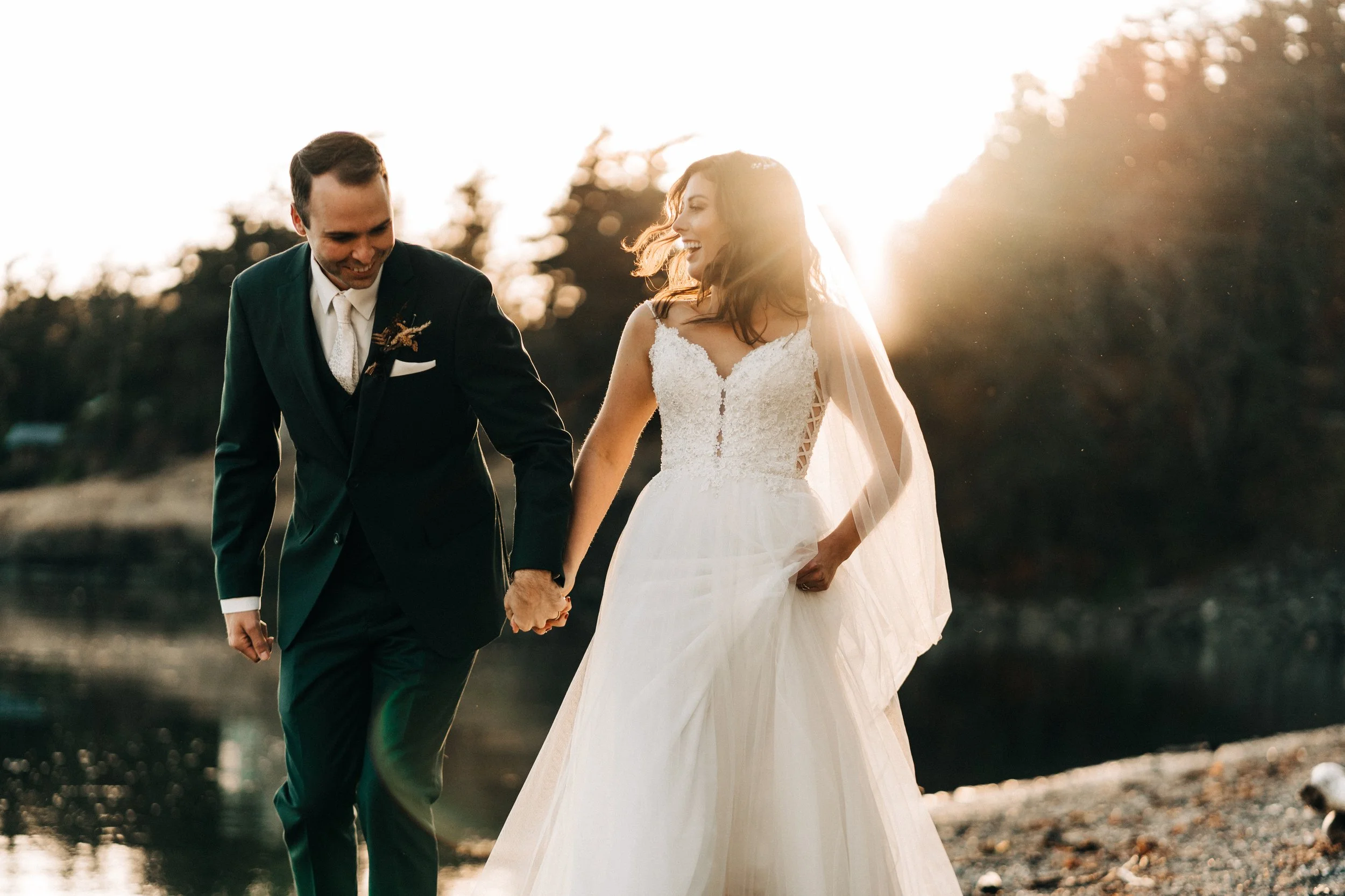 A newlywed couple holding hands on a rocky beach during sunset, smiling and looking at each other.