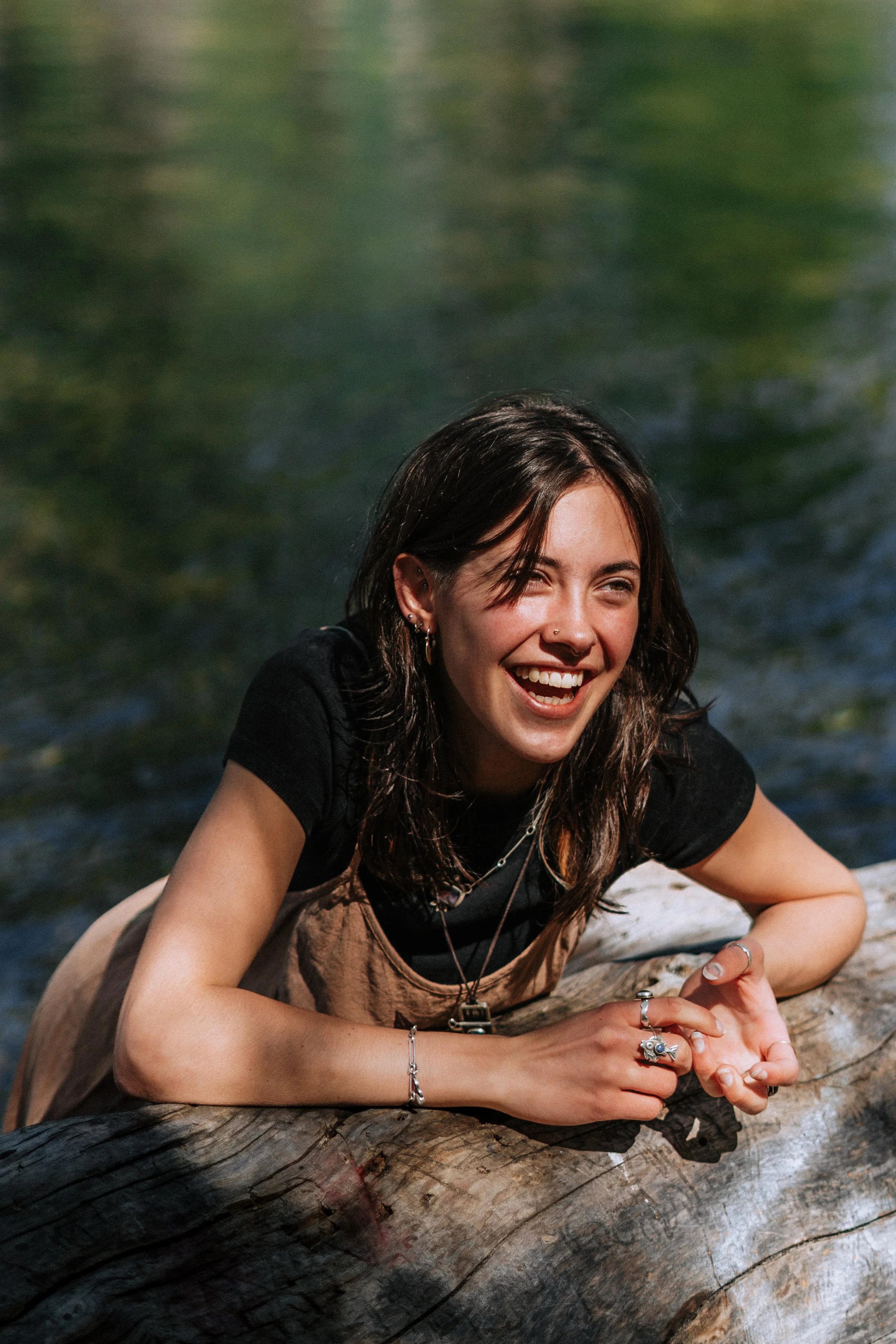 A young woman with dark hair and earrings, smiling and laughing, lying on her stomach on a log near a body of water.