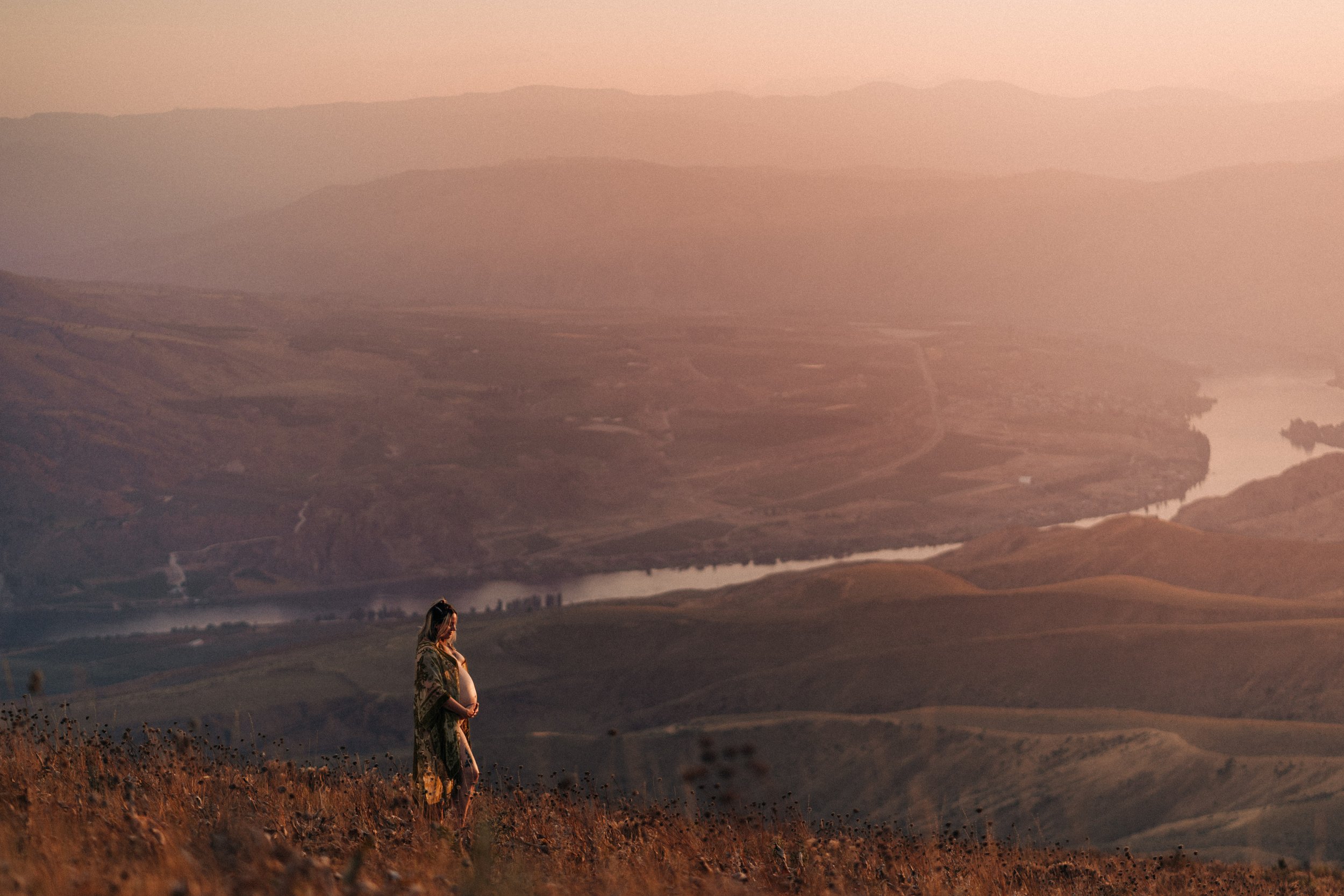 Person standing on hillside overlooking a valley with a river at sunset.