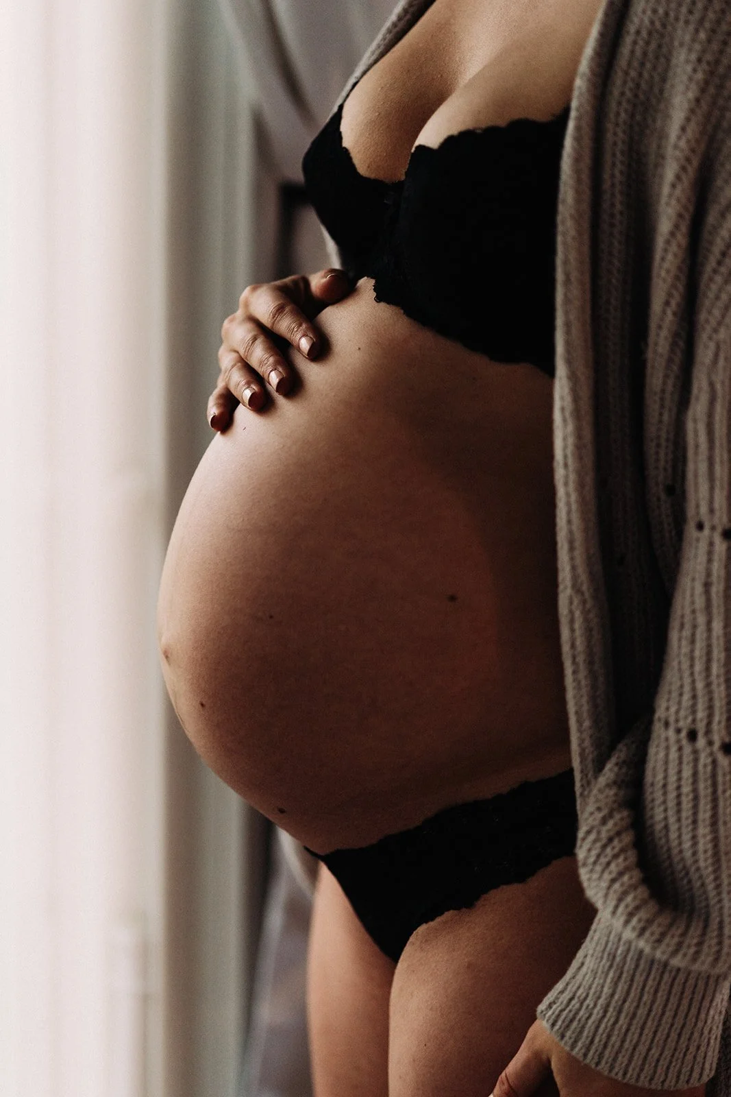 Close-up of a pregnant woman standing near a window, gently touching her belly with one hand. She is wearing black lace underwear and a striped cardigan | Chelan Maternity Photographer