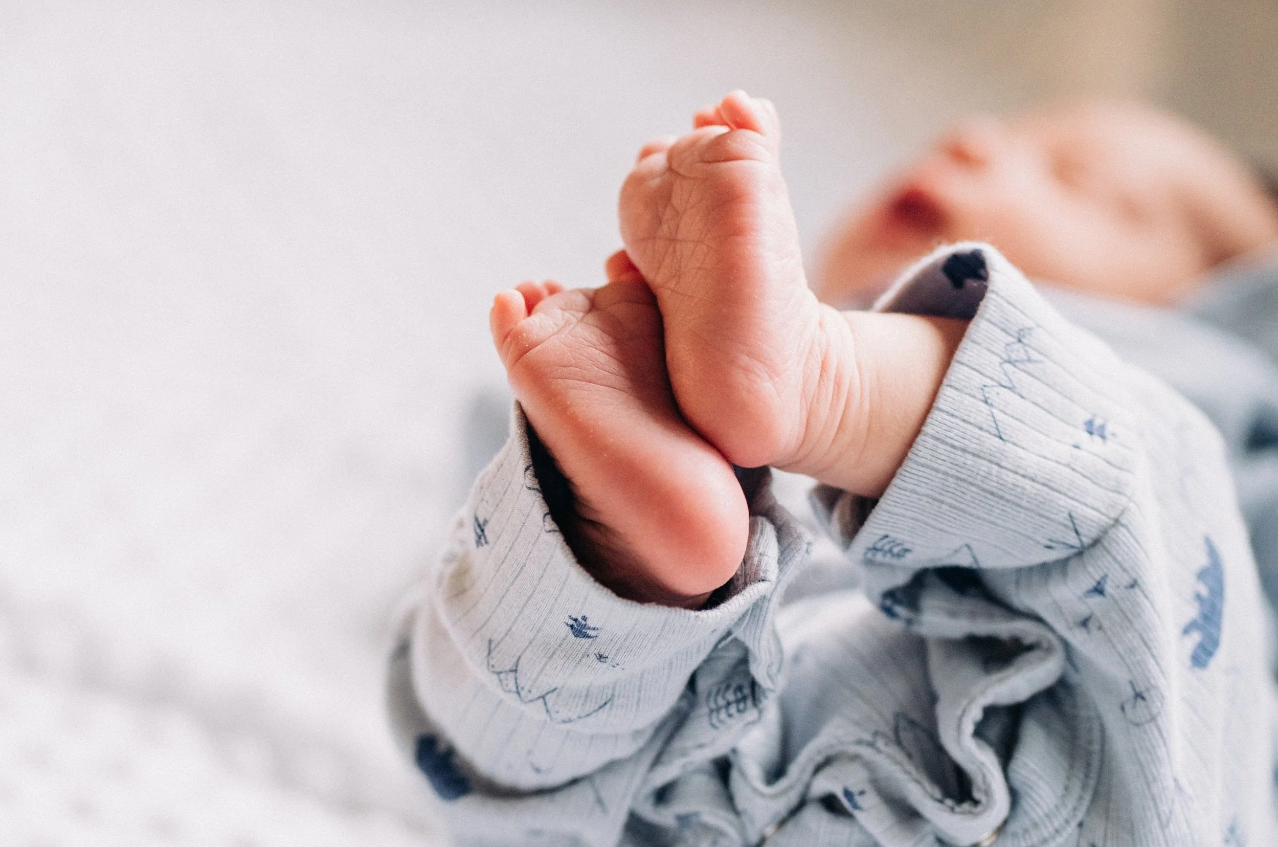 Close-up of a newborn baby holding a finger with both hands, wearing a light-colored onesie with small blue and gray patterns | Chelan Newborn Photographer