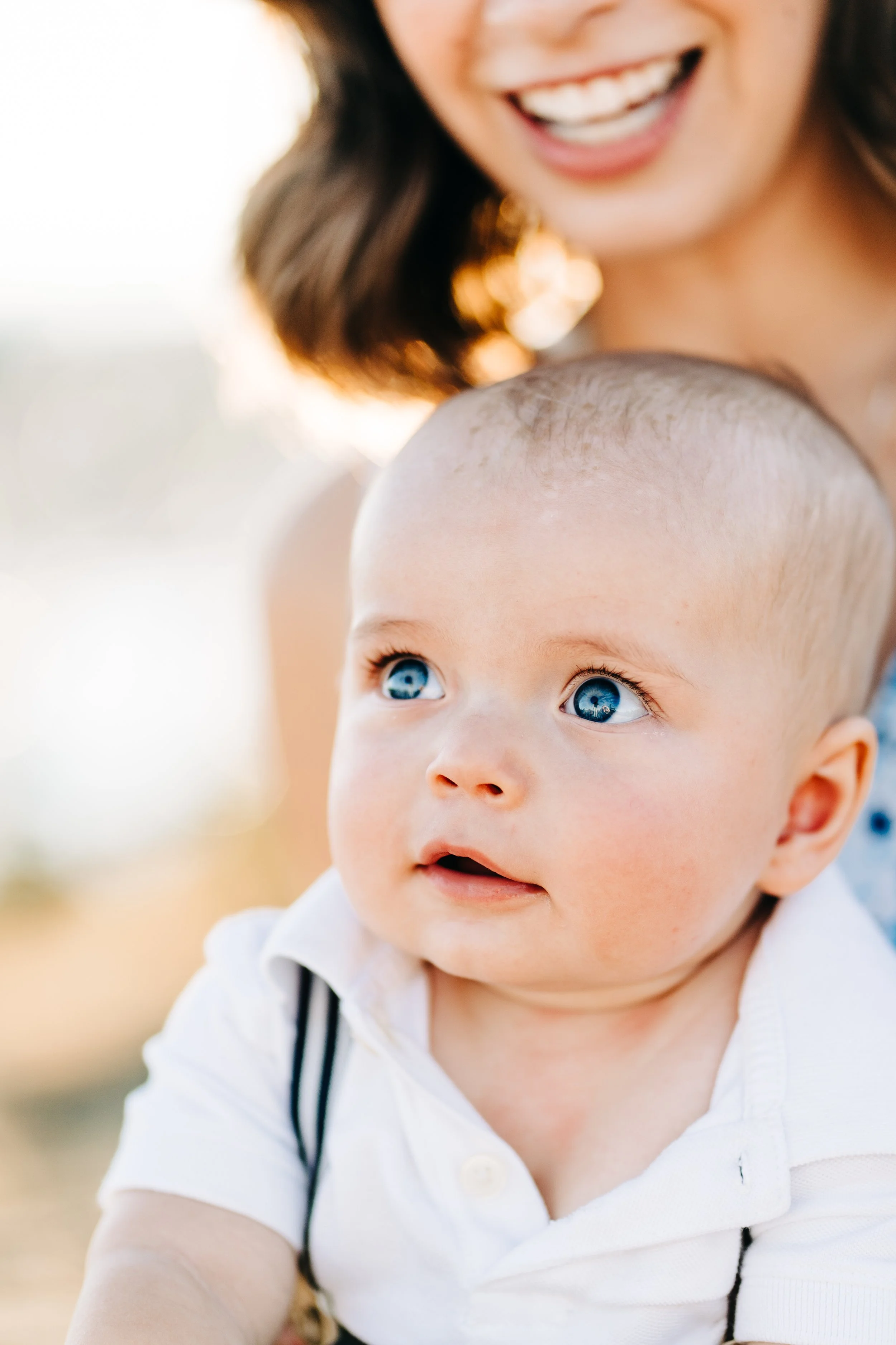 Close-up of a smiling woman with brown hair holding a young boy with bright blue eyes and a pacifier, outdoors during daytime.