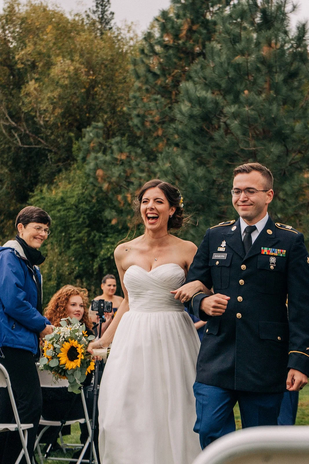 A woman in a white wedding dress walking arm-in-arm with a man in a military uniform at an outdoor wedding ceremony, with guests and trees in the background.
