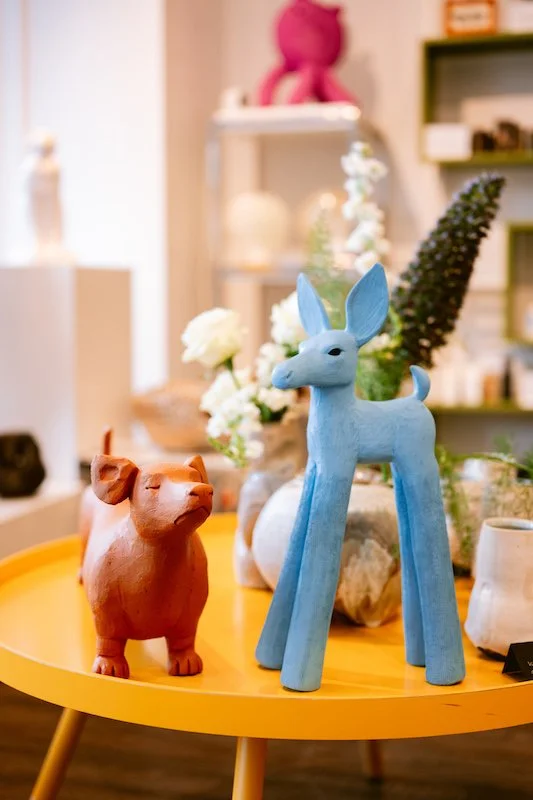 Aritst-made ceramic animal sculptures of a blue deer and a brown dachshund dog, on a yellow table with vases of white and purple flowers in the background.
