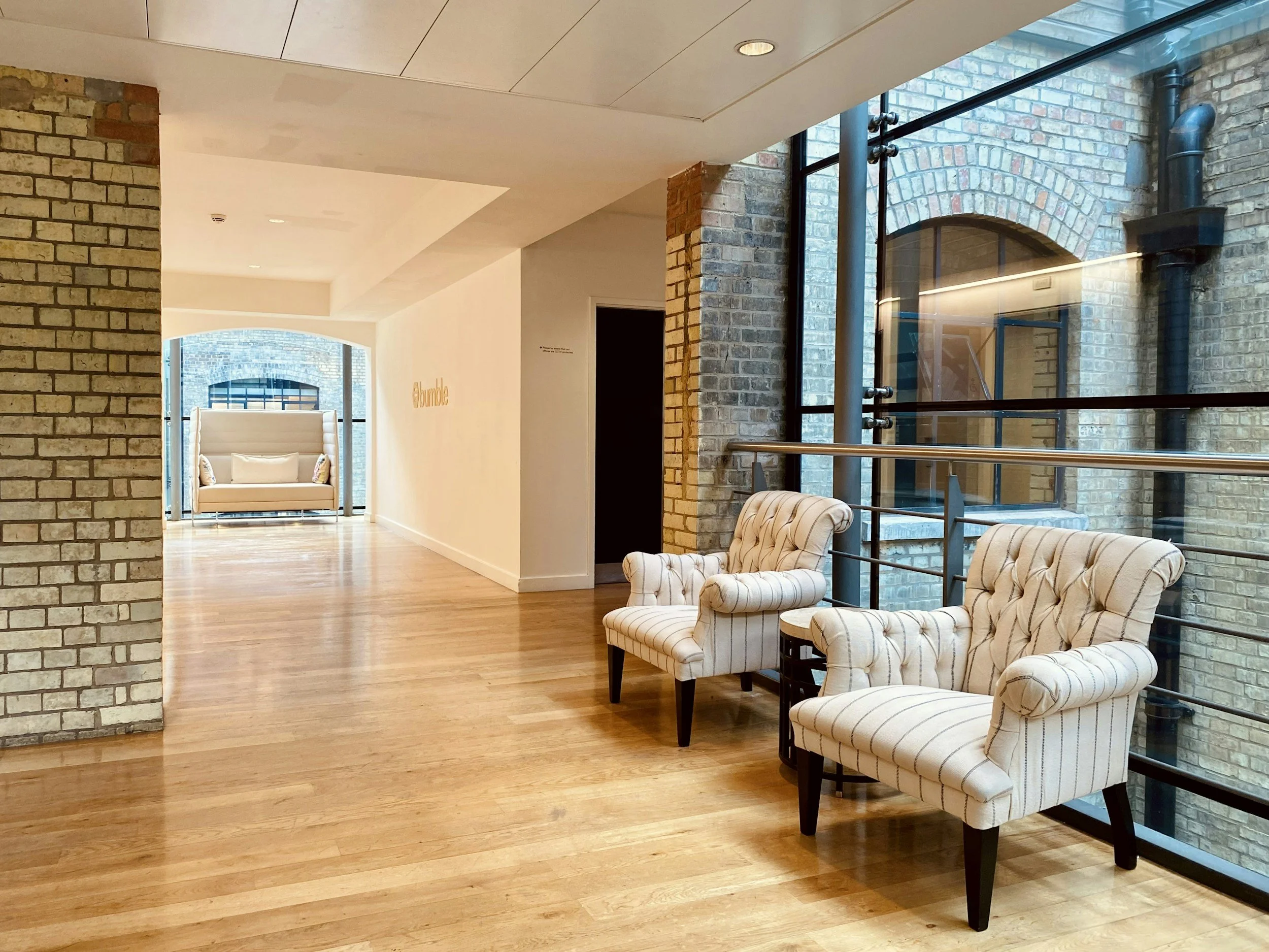 Interior of a modern building with exposed brick walls, hardwood floors, large windows, and striped armchairs.
