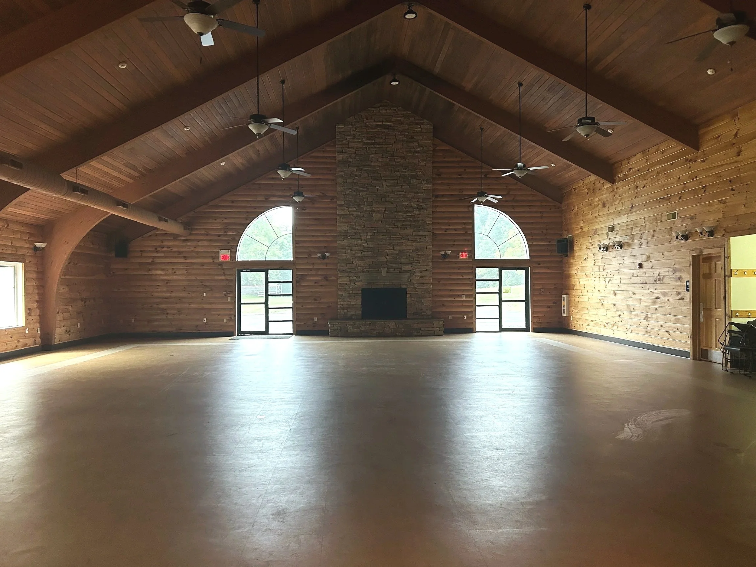 A large, open room with wooden walls and ceiling, multiple ceiling fans, a stone fireplace in the center, large arched windows, and double glass doors.