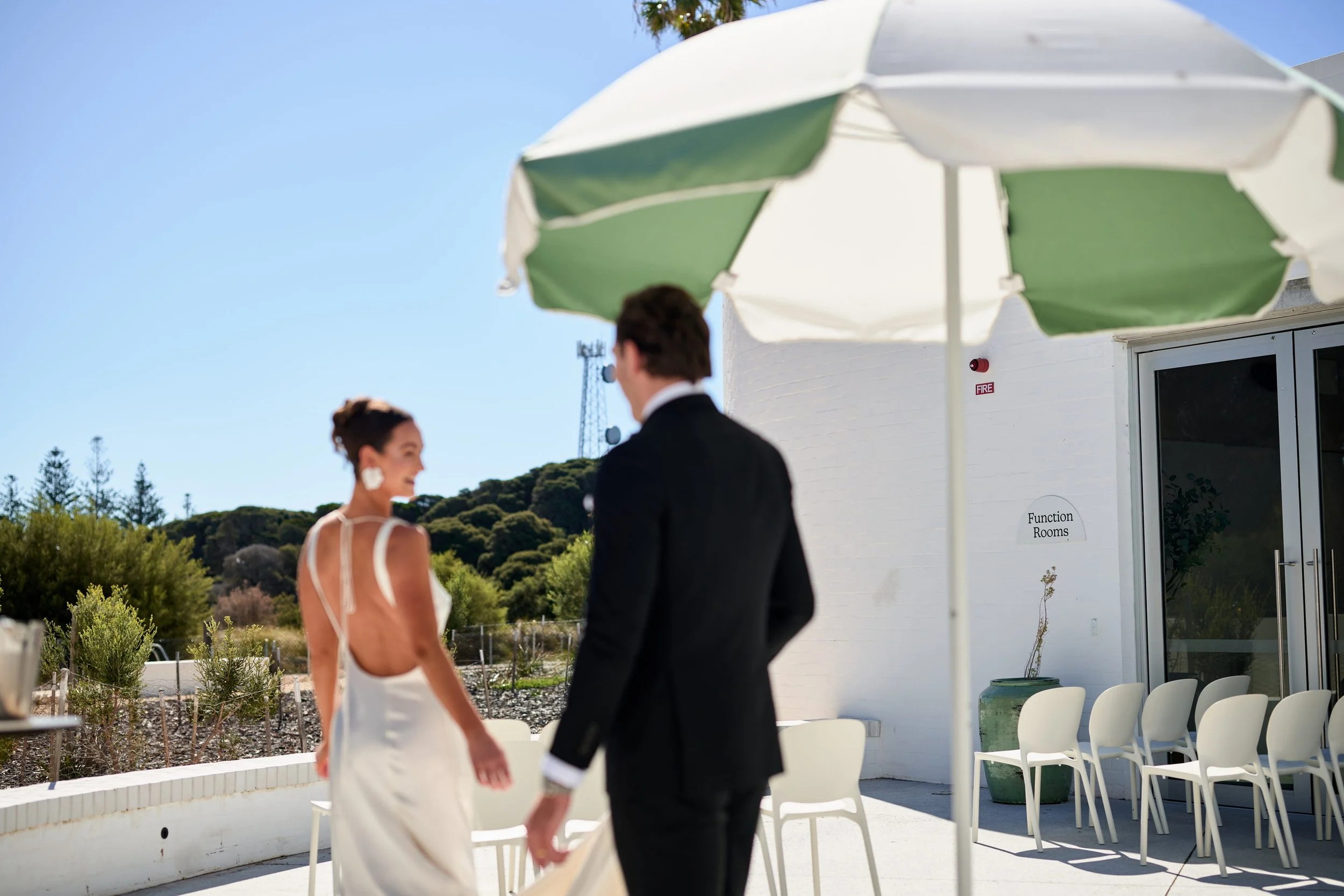 A couple walking across a sunlit terrace beside a green and white umbrella
