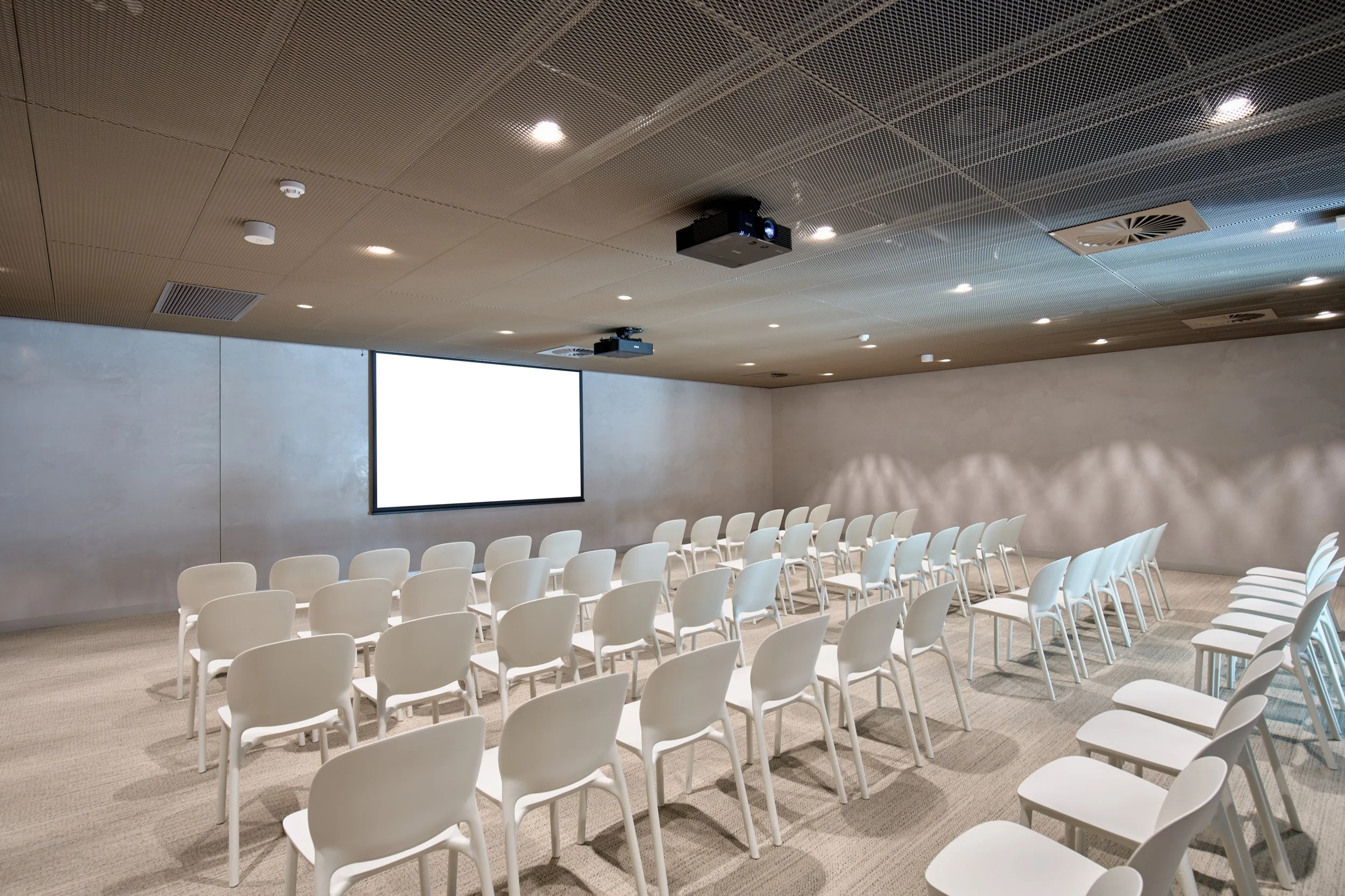 Empty conference room with white chairs arranged in rows facing a large white projection screen on the wall, ceiling-mounted projector, and neutral-toned walls and carpeted floor.