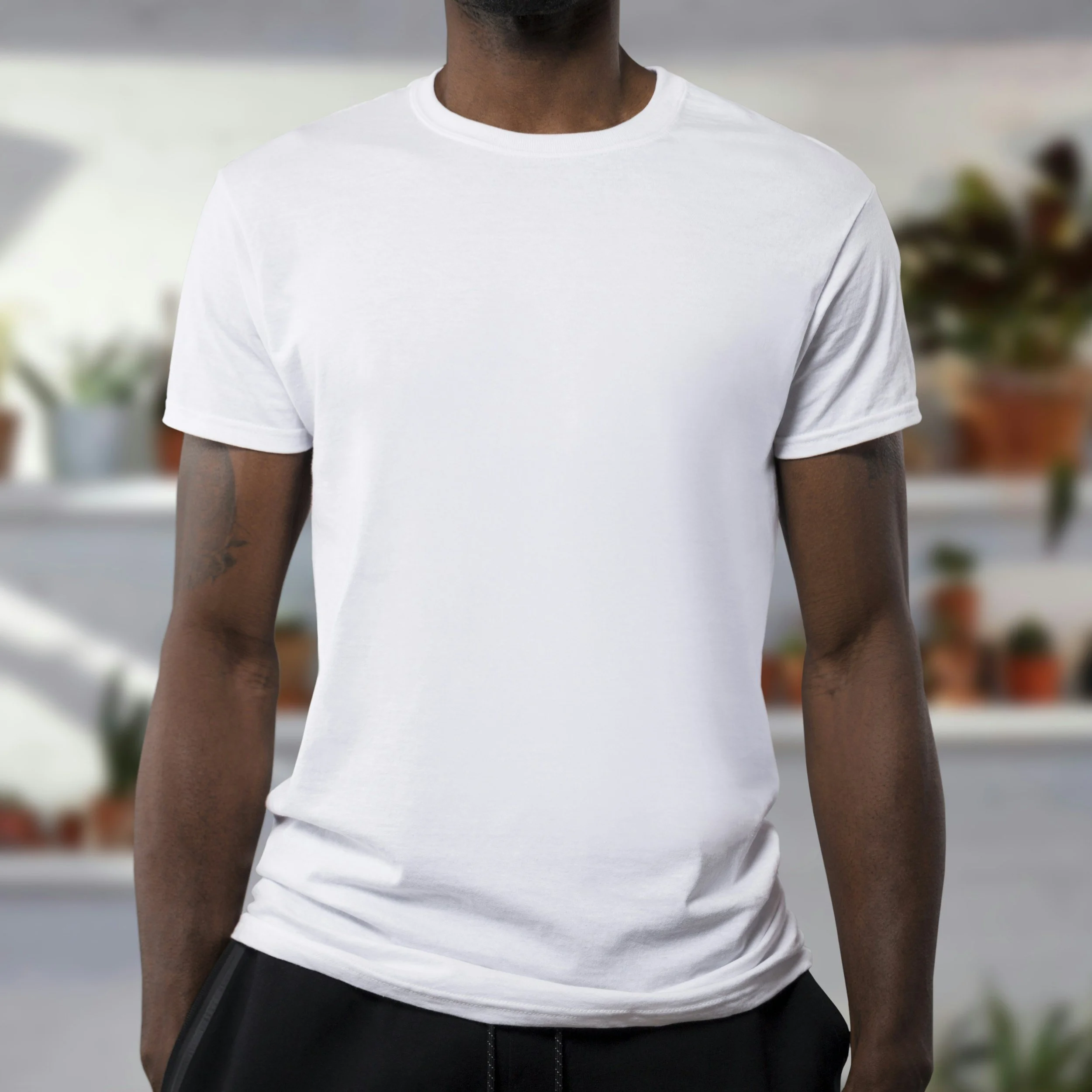 Front view of a man wearing a plain white T-shirt, standing indoors with a blurred background of potted plants.
