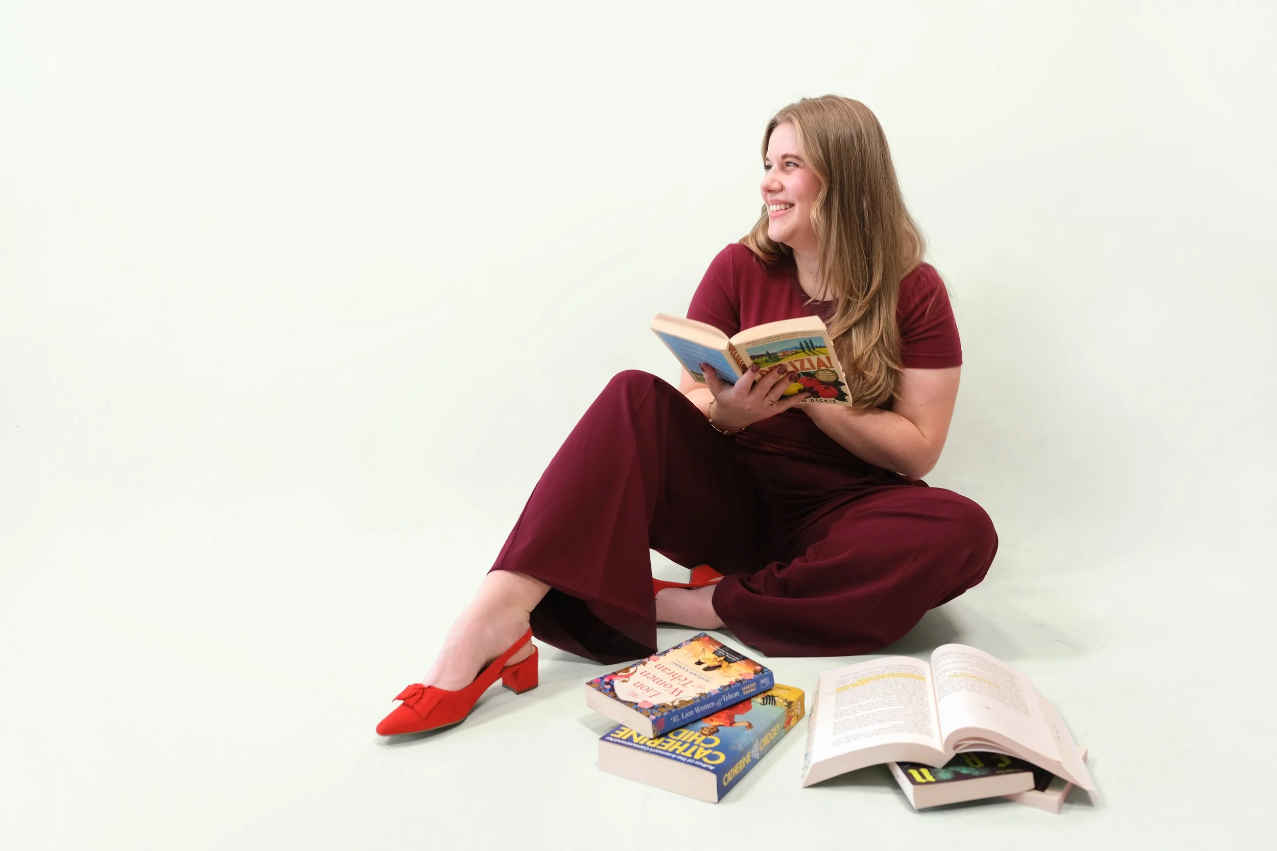 A woman is sitting on the ground, holding a book, there are books on the floor in front of her