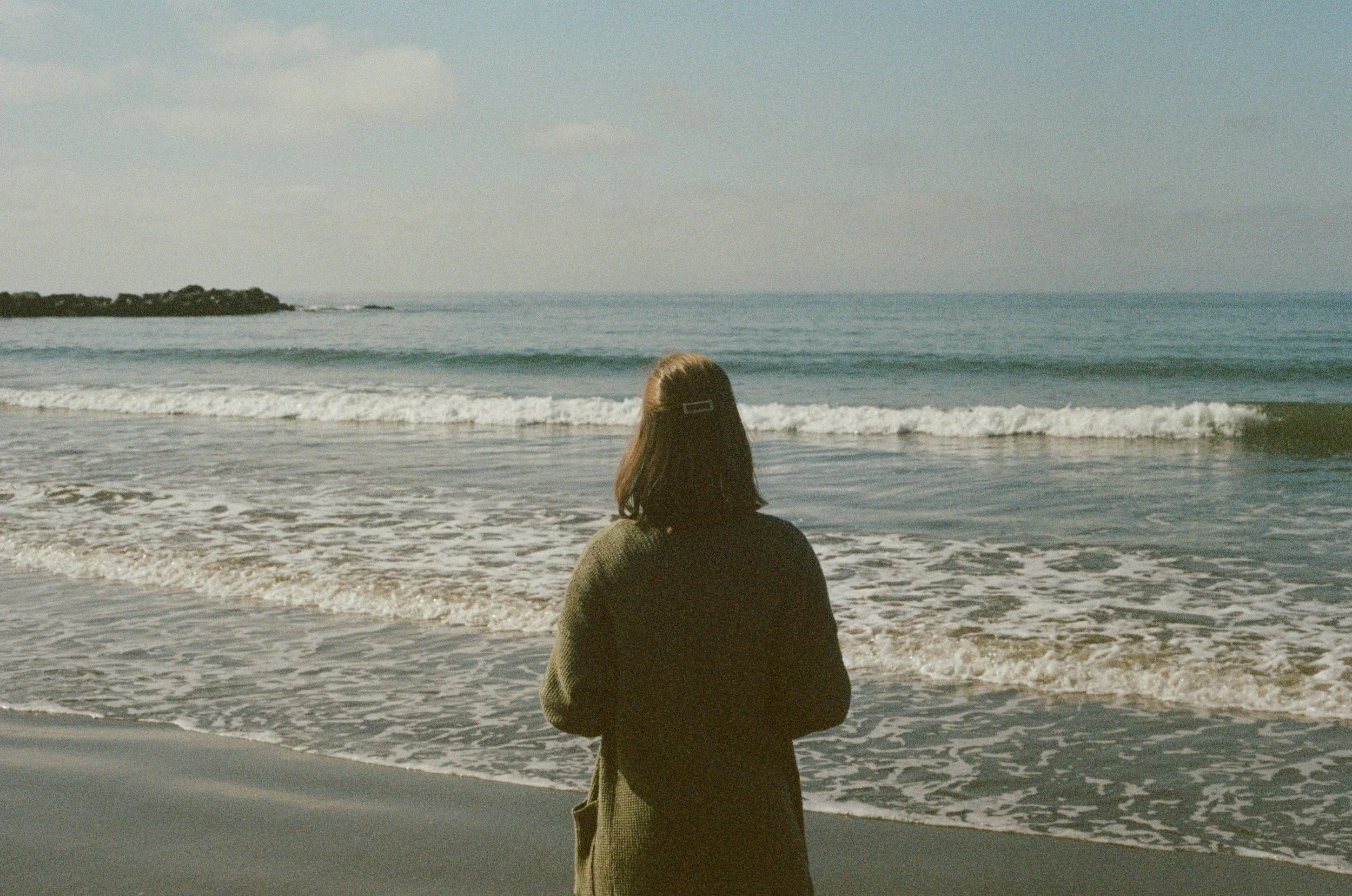 Woman with shoulder length brown hair looking off into ocean with waves rolling in.