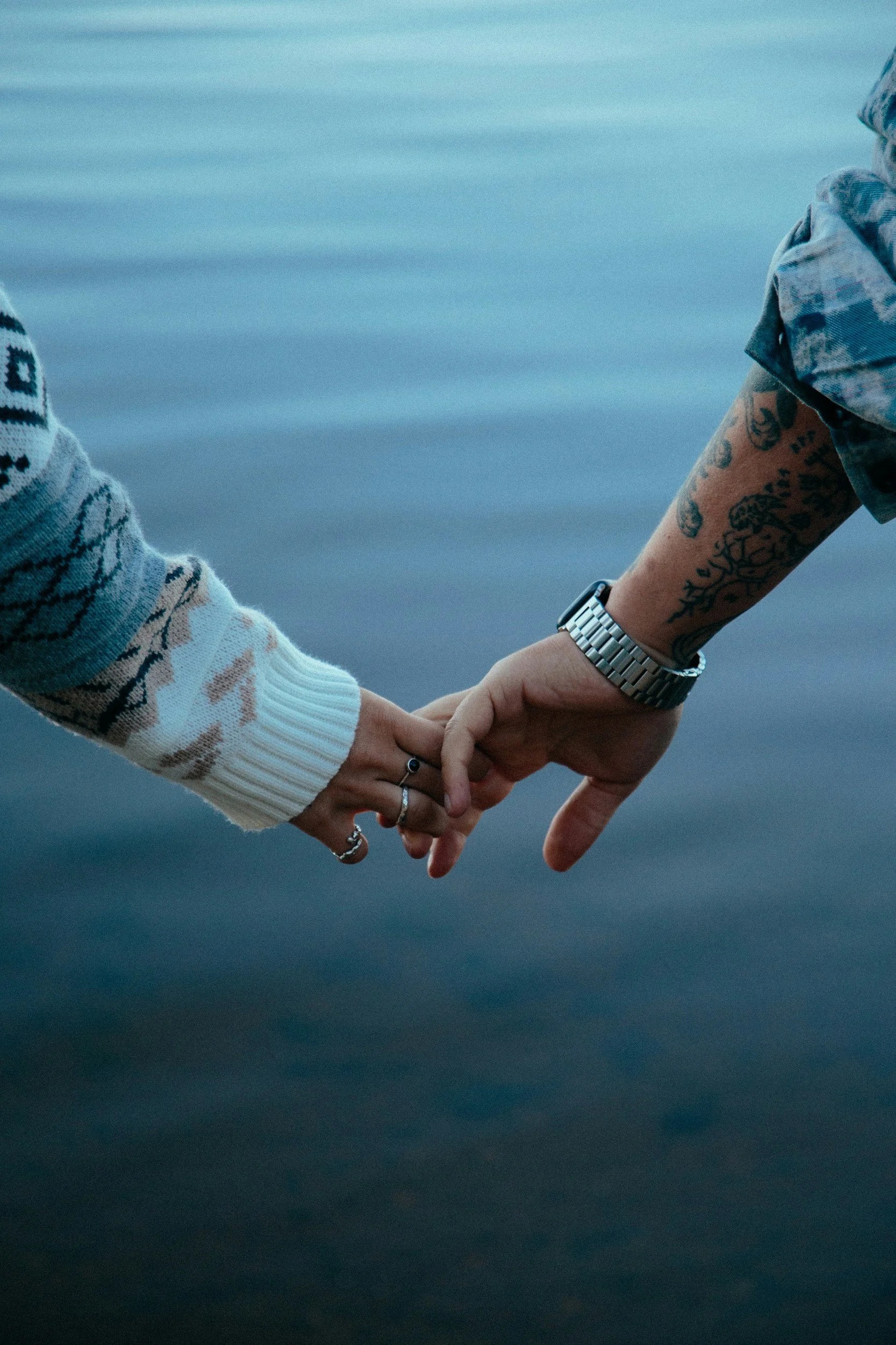 Couple holding hands with only hands in shot, one partner wearing rings, the other a watch with tattoos up the forearm.
