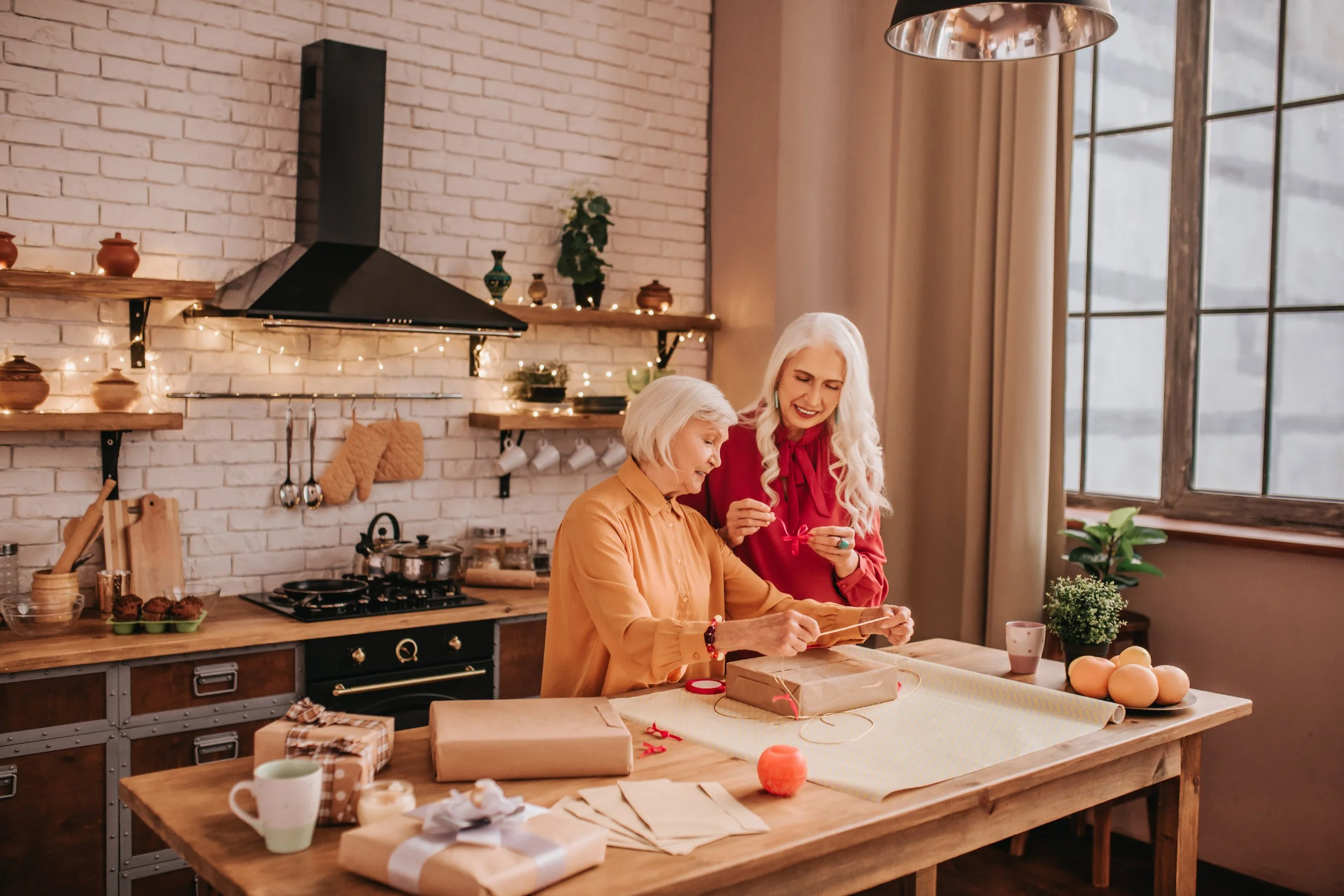 Two women, an elderly woman and a middle-aged woman, are wrapping gifts together in a cozy kitchen decorated for Christmas, with a brick wall, a large window, and various kitchen items and plants around them.