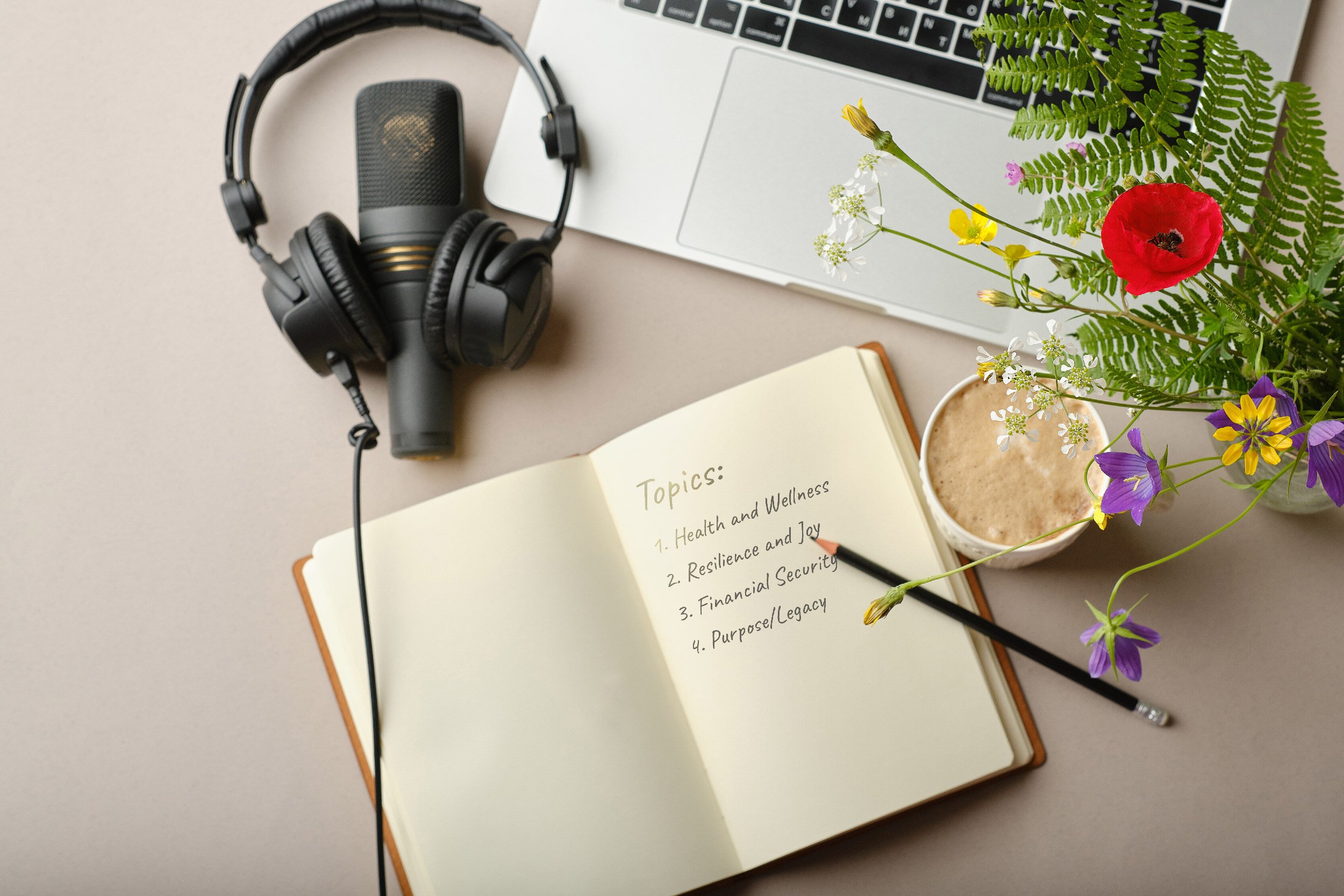 A workspace with a notebook open to a list of topics including health, resilience, financial security, and purpose; a pair of headphones, a laptop, a cup of coffee, and a vase of colorful flowers.