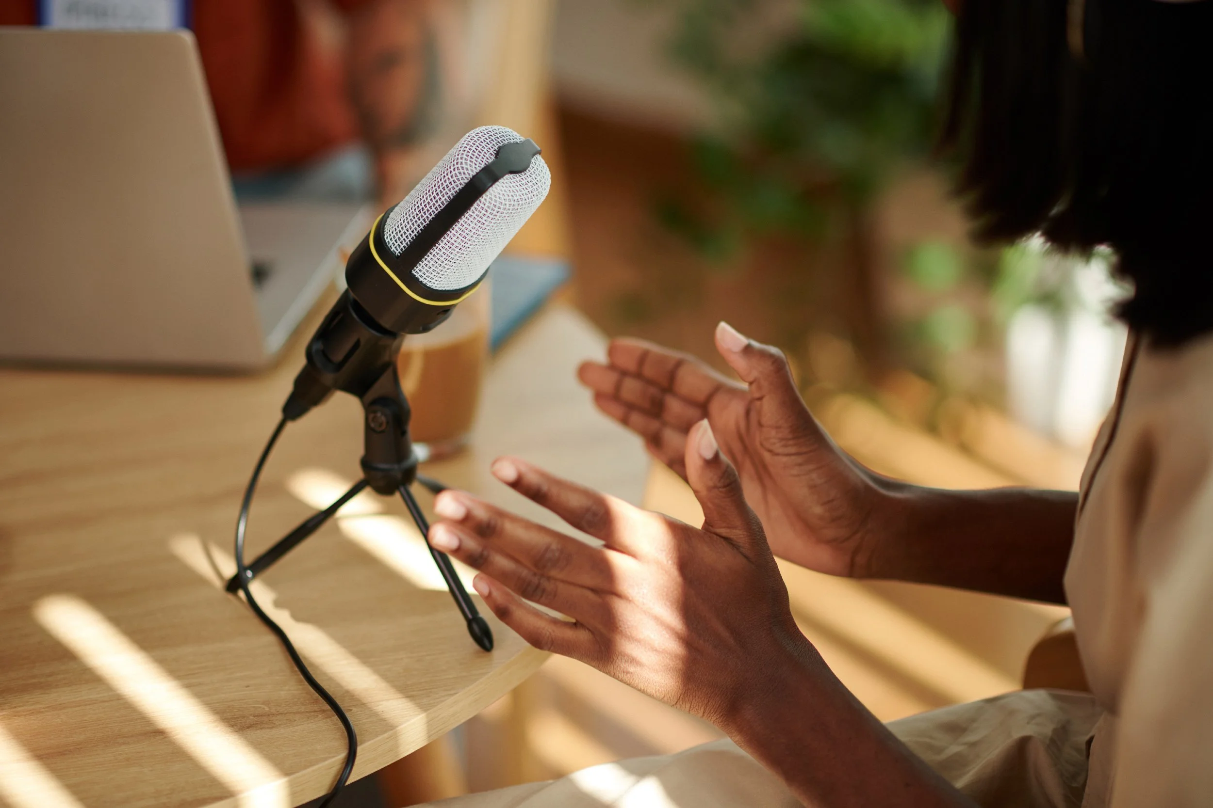 Person gesturing with hands in front of a microphone on a desk, with a laptop in the background.