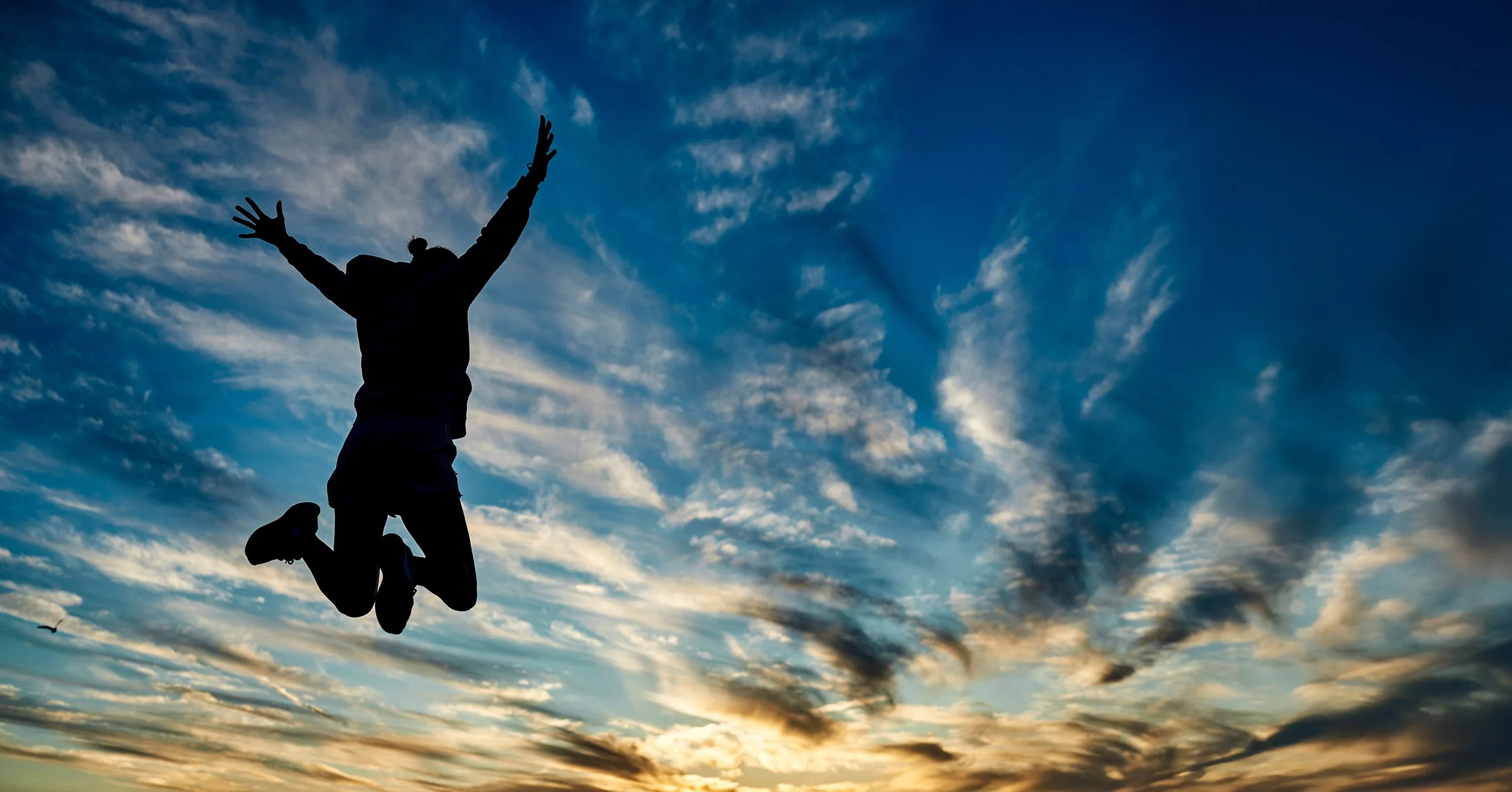 Person jumping in the air at sunset with a dramatic sky and clouds in the background.