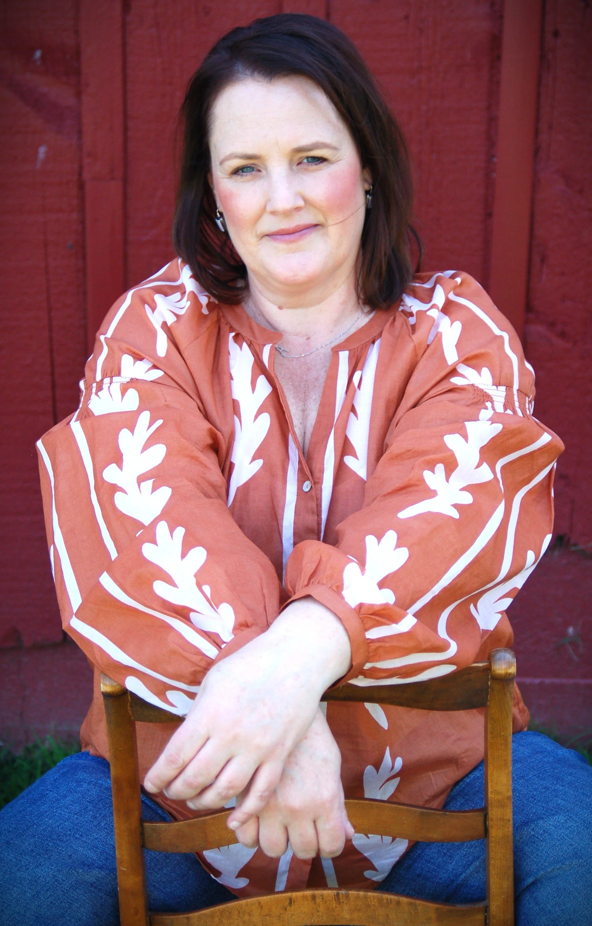 Woman with dark hair wearing an orange and white patterned shirt, sitting in front of a red wooden wall.