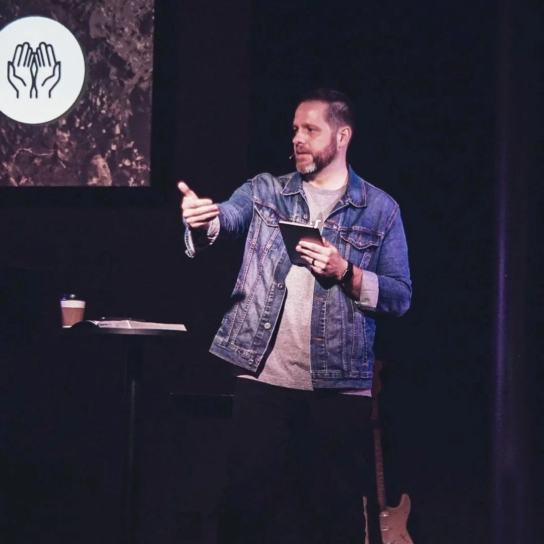A man in a denim jacket giving a presentation on stage with a slide showing two hands in a caring gesture and a coffee cup nearby.