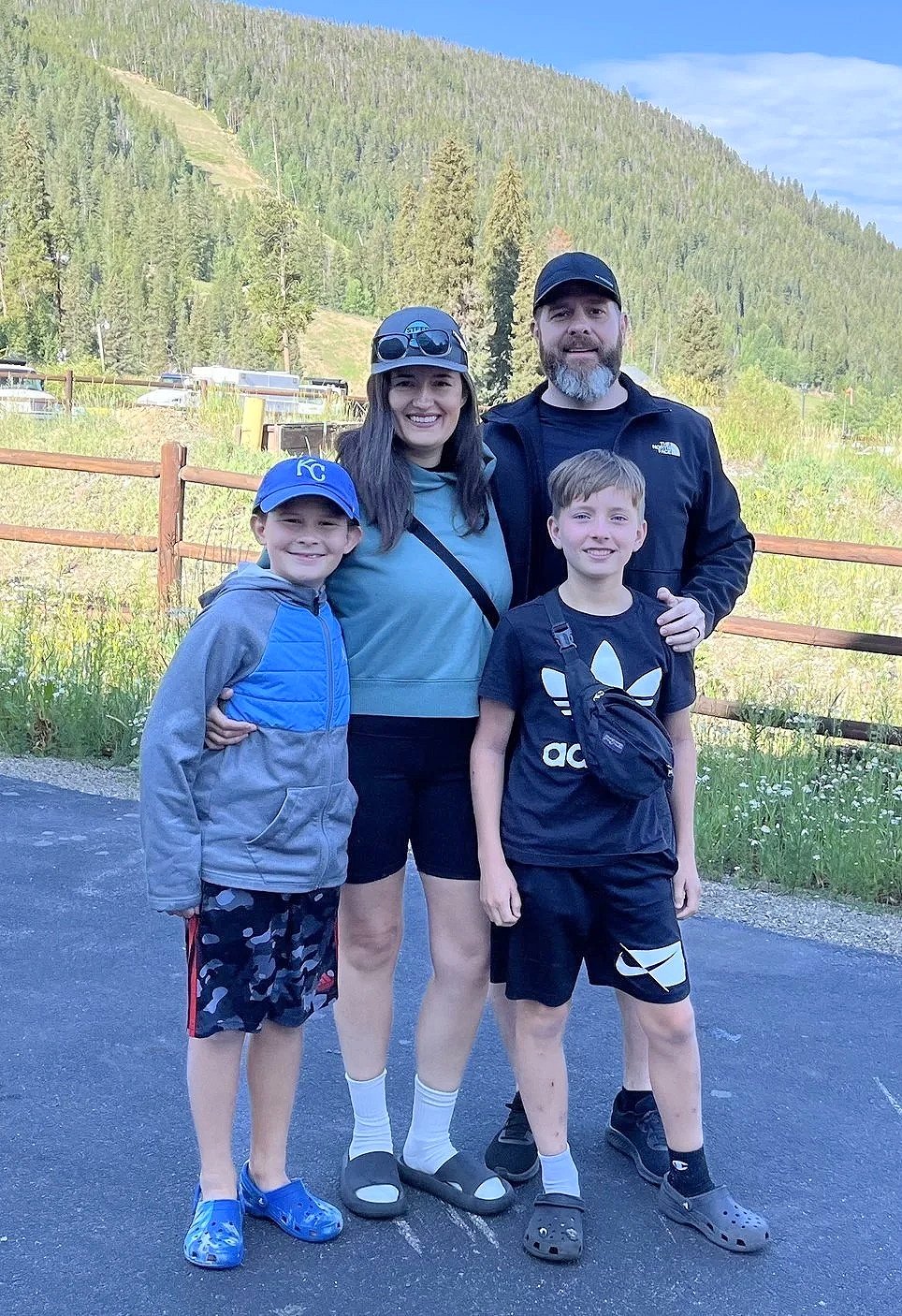 A family of four standing outdoors on a paved path with green mountains and trees in the background. The woman is wearing a baseball cap and sunglasses, the man is wearing a cap, and two boys are smiling in front, one wearing a Kansas City Royals cap and the other wearing an Adidas shirt.