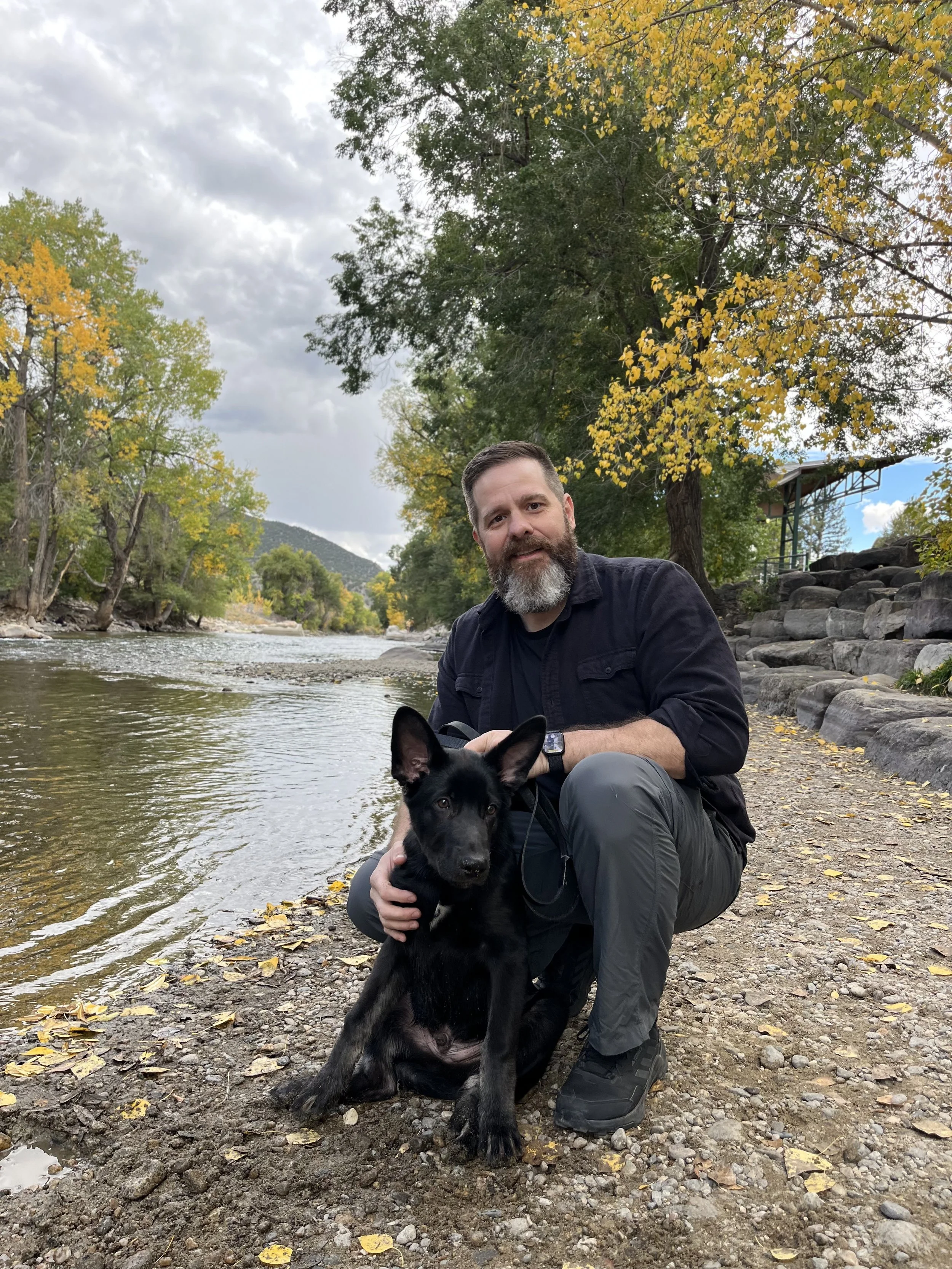 A man with a beard in outdoor clothing kneeling next to a black and tan dog near a riverbank with rocks and fallen leaves, surrounded by trees with autumn foliage, under a cloudy sky.