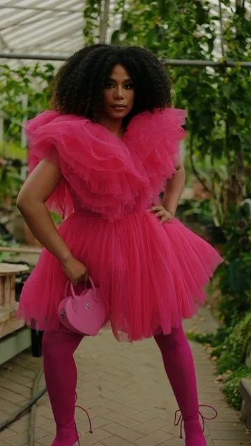 Woman in a vibrant pink ruffled dress with matching pink tights holding a pink purse, standing in a greenhouse surrounded by green plants.
