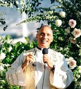 A man dressed in white religious attire holding a microphone and giving a speech outdoors, surrounded by green foliage and white flowers.