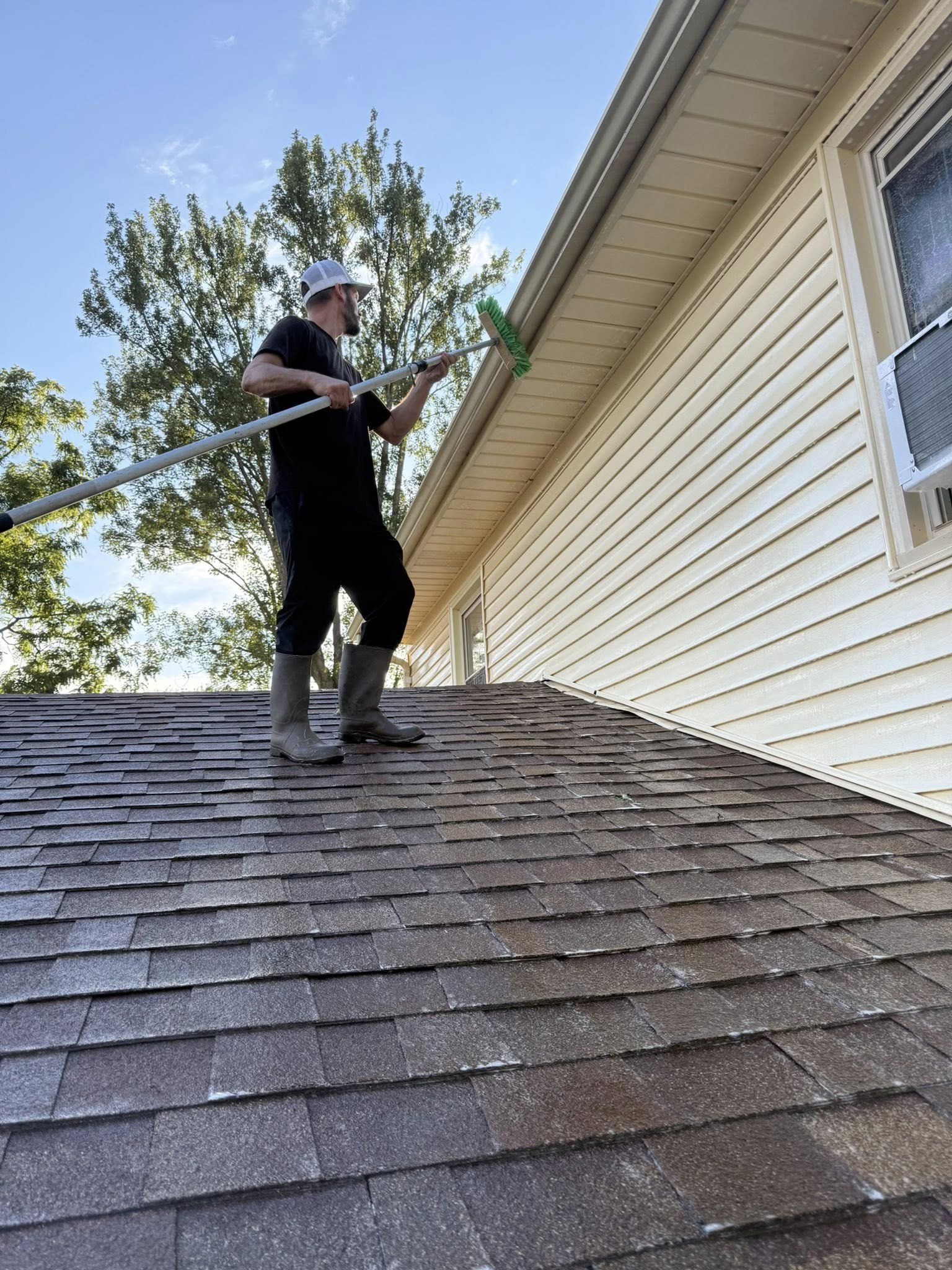 A man wearing rubber boots, a black t-shirt, and a white cap is standing on a roof and cleaning the exterior wall of a house with a long-handled cleaning tool.