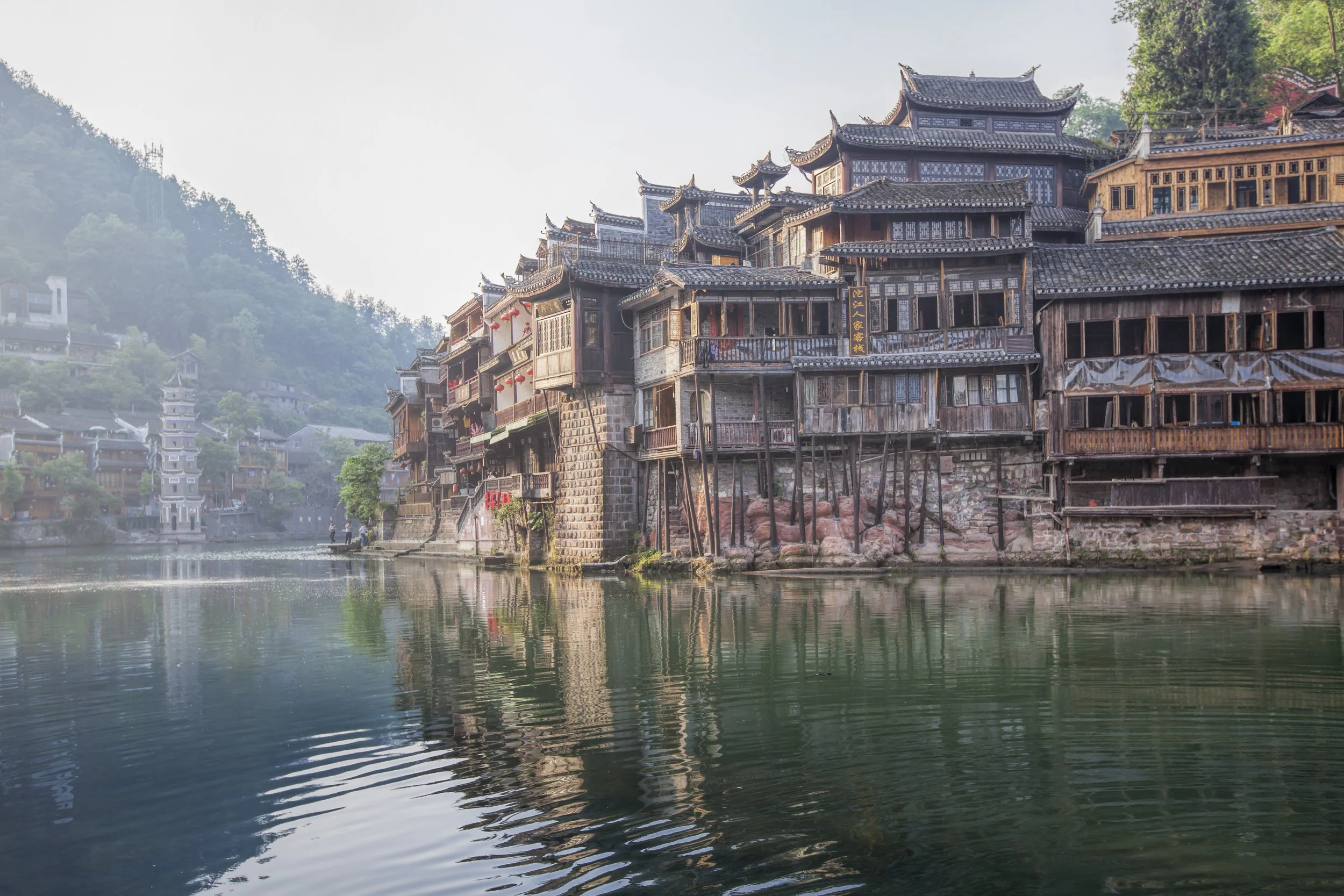 Traditional wooden Chinese buildings along a river, with a hillside and trees in the background.