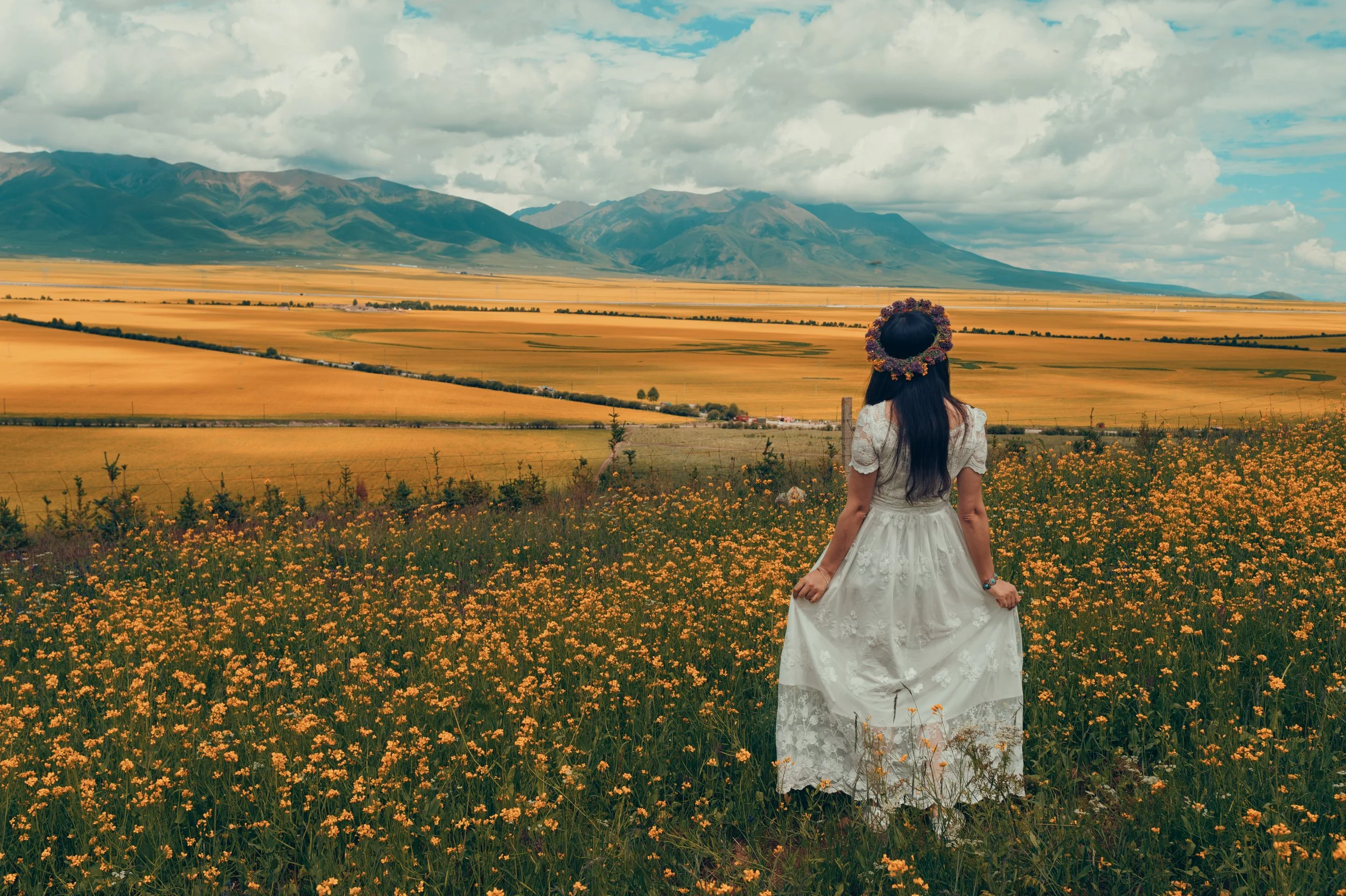 Woman dressed in a white lace dress with a floral crown standing in a field of yellow flowers, facing a wide landscape of farmland and mountains under a partly cloudy sky.