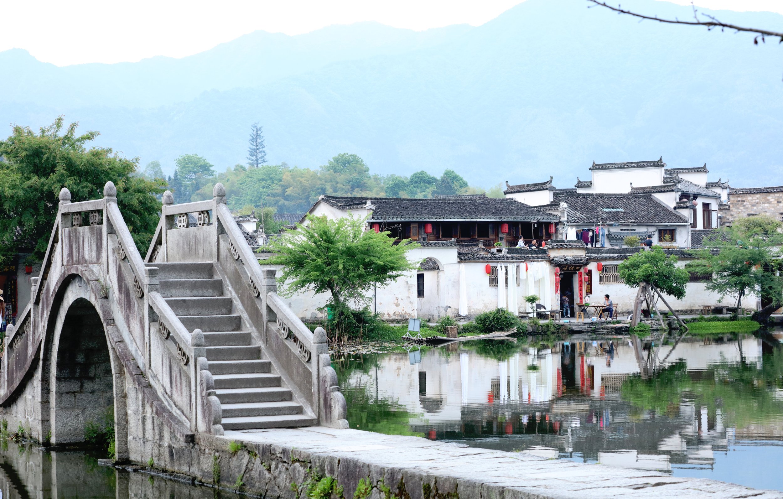 Traditional Chinese white-walled houses with dark tiled roofs along a waterway, featuring a stone arched bridge, lush green trees, and mountains in the background.