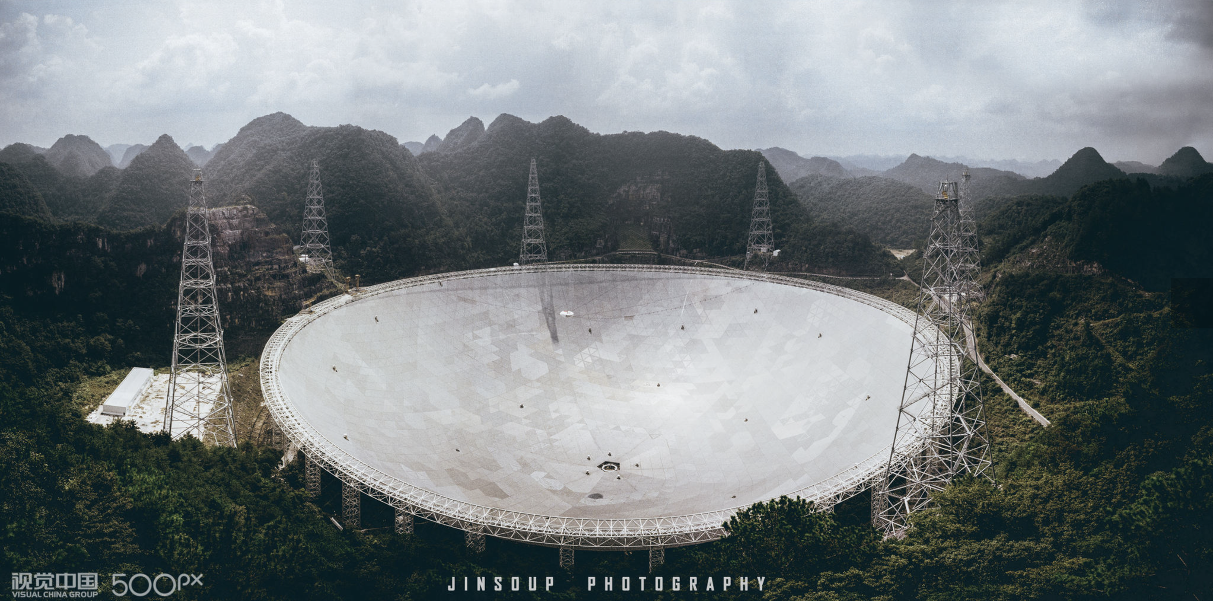 Large satellite dish surrounded by green hills and mountains with cloudy sky.
