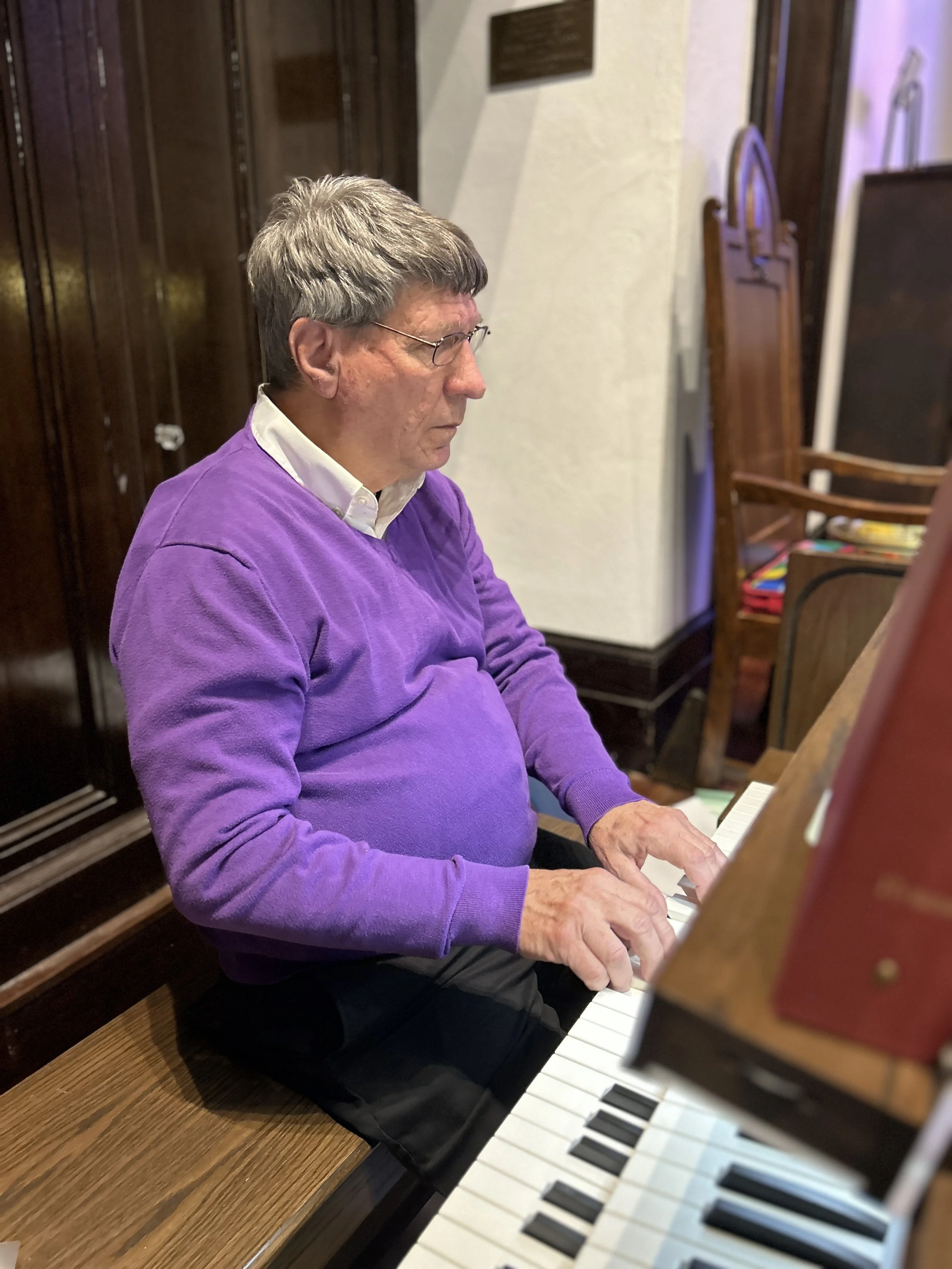 A man with curly dark hair, glasses, and a beard, wearing a dark green sweater, sitting indoors against a blurred background with a plant and wall decor.