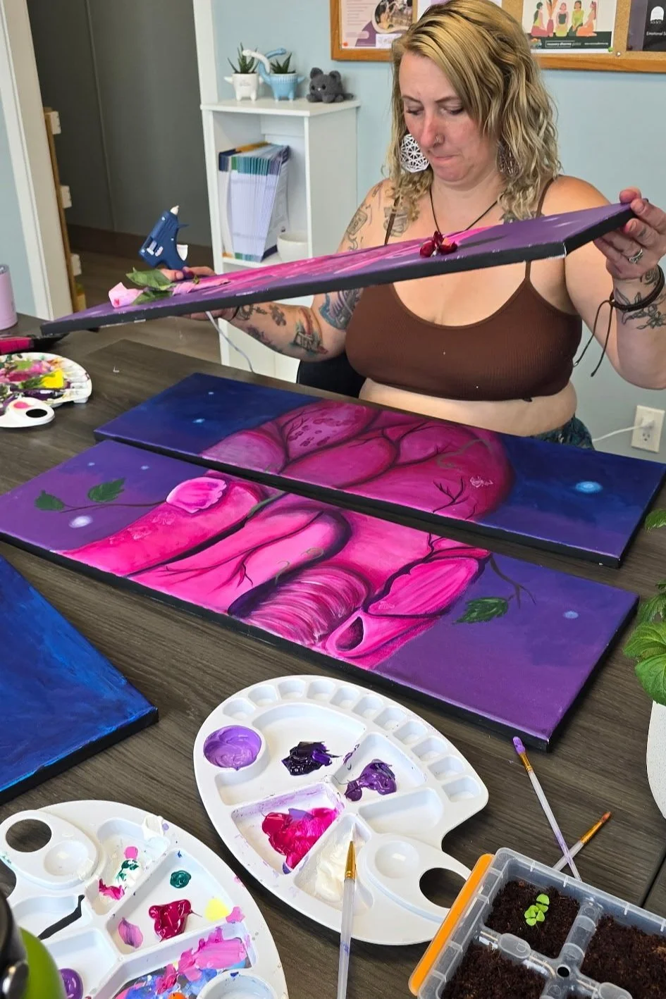 Woman in a brown tank top painting a pink and purple abstract artwork featuring a tree and leaves on a canvas in an art studio.