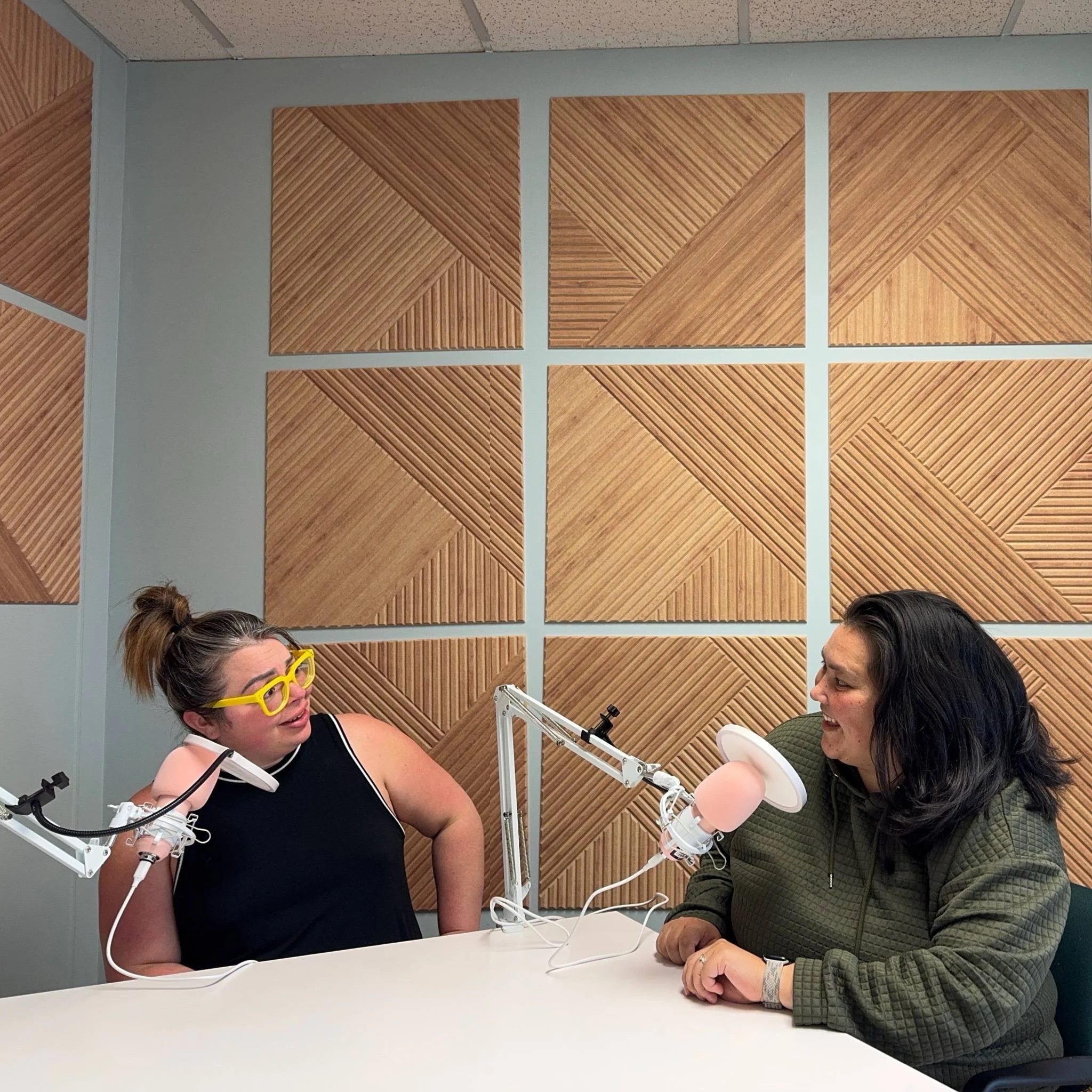 Two women sit at a table in a recording studio, smiling and talking to each other. The woman on the left has her hair in a bun, wears yellow glasses and a black sleeveless top. The woman on the right has dark hair, wears a green jacket, and is speaki