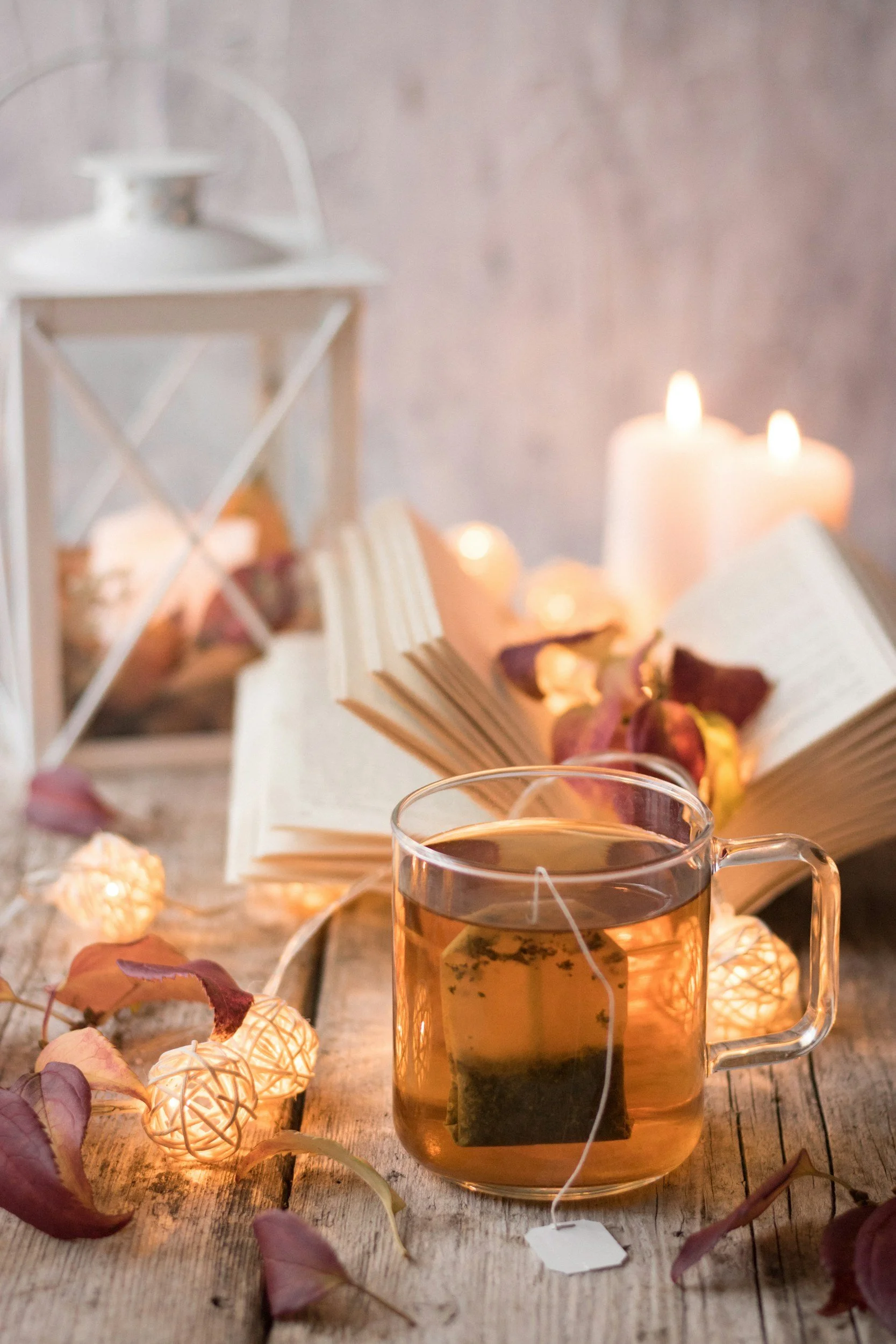 A mug of hot tea on a rustic wooden table surrounded by autumn leaves, decorative string lights, an open book, and lit candles, creating a cozy ambiance.