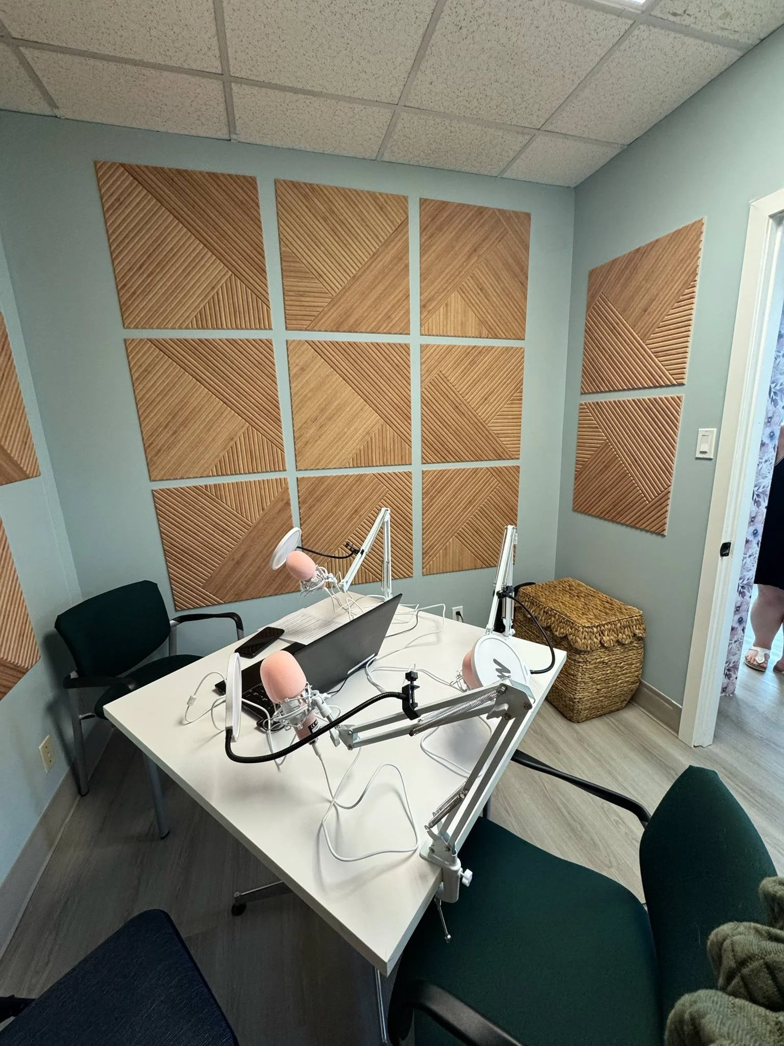 An empty recording studio with a white table, three microphones with pop filters, a laptop, and two chairs, one black and one green, in a room with acoustic wooden panels on the wall and a wicker basket in the corner.