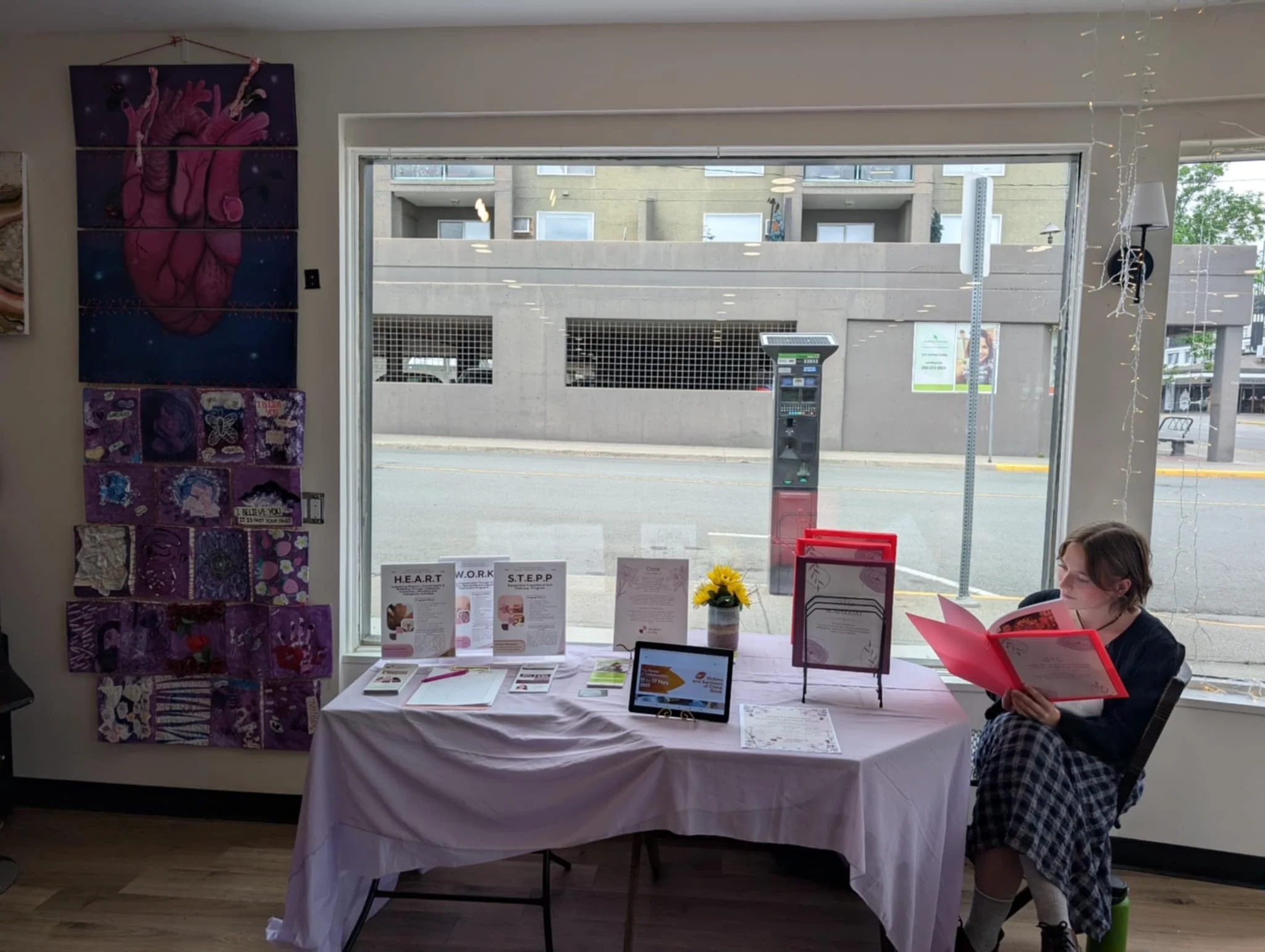 Girl sitting by a table with brochures, signs, and a potted sunflower, next to a large window with a view of a parking lot and street outside.