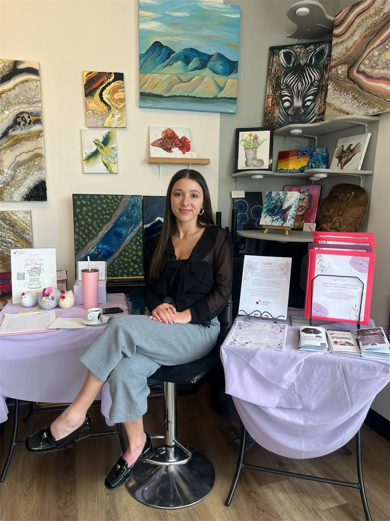 A young woman sitting at a table in an art gallery or store, surrounded by various paintings and artworks on the wall and shelves, with informational pamphlets and decorative items on the tables.