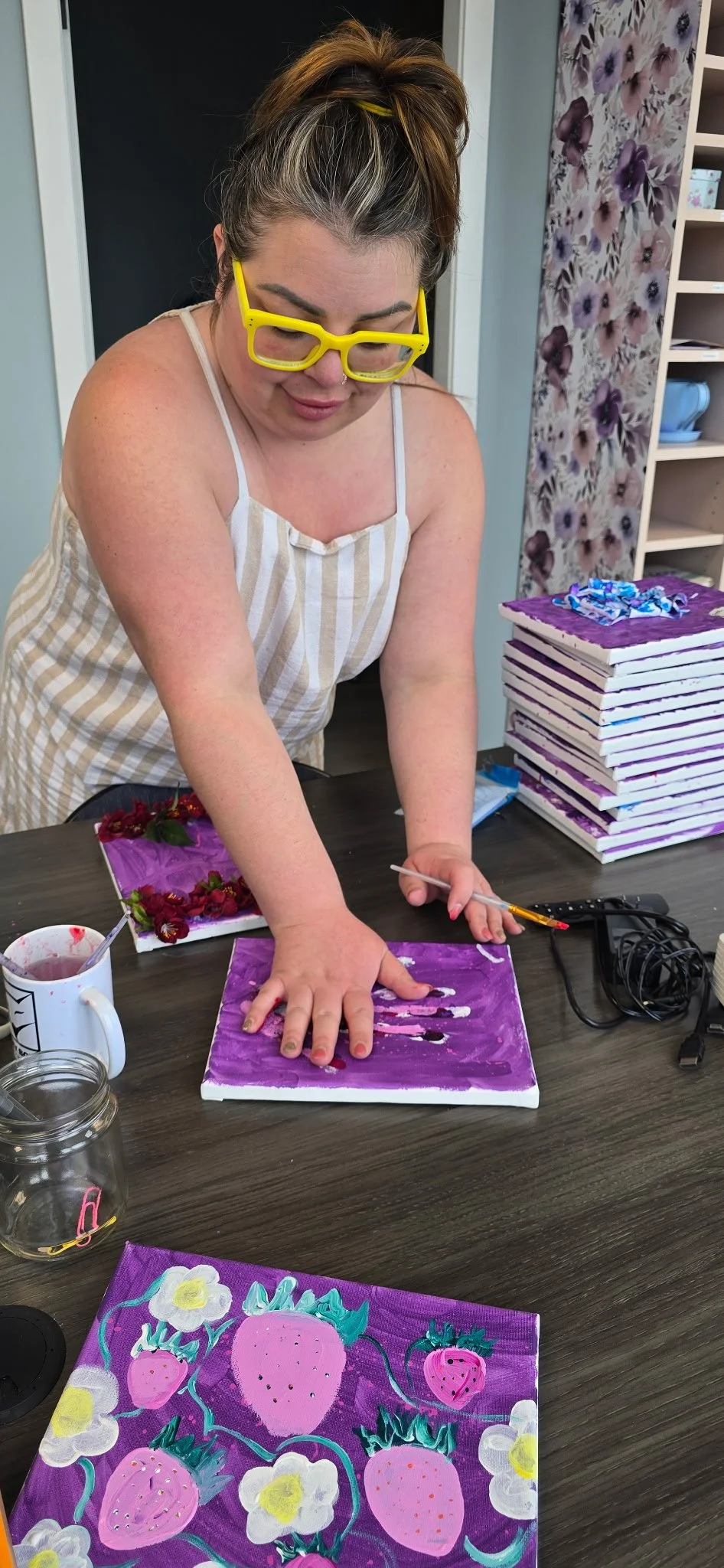 Woman wearing yellow glasses creating a mixed media art piece with purple background and textured pink strawberries and white flowers. She is pressing down on canvas with her hand, surrounded by art supplies and stacked canvas boards.