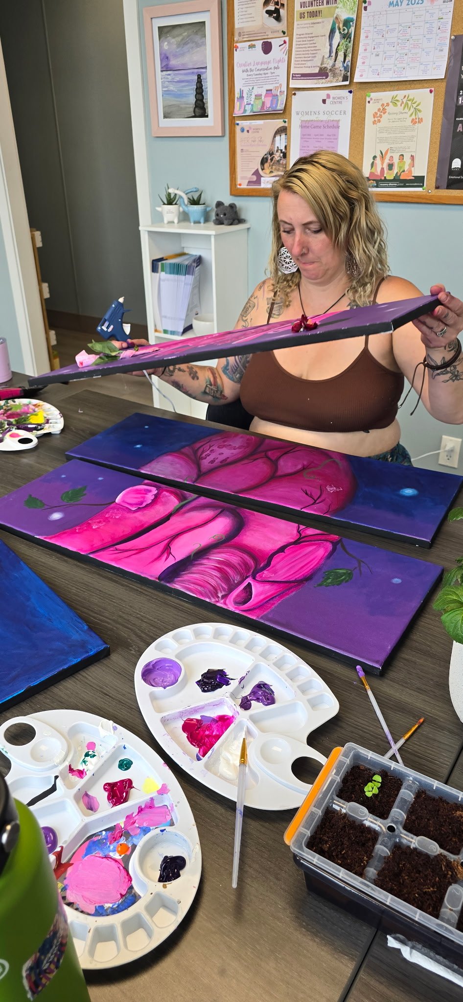 Woman painting a pink tree on a canvas with a purple night sky background, sitting at a table with painting supplies in a cozy room with bulletin boards and decor.