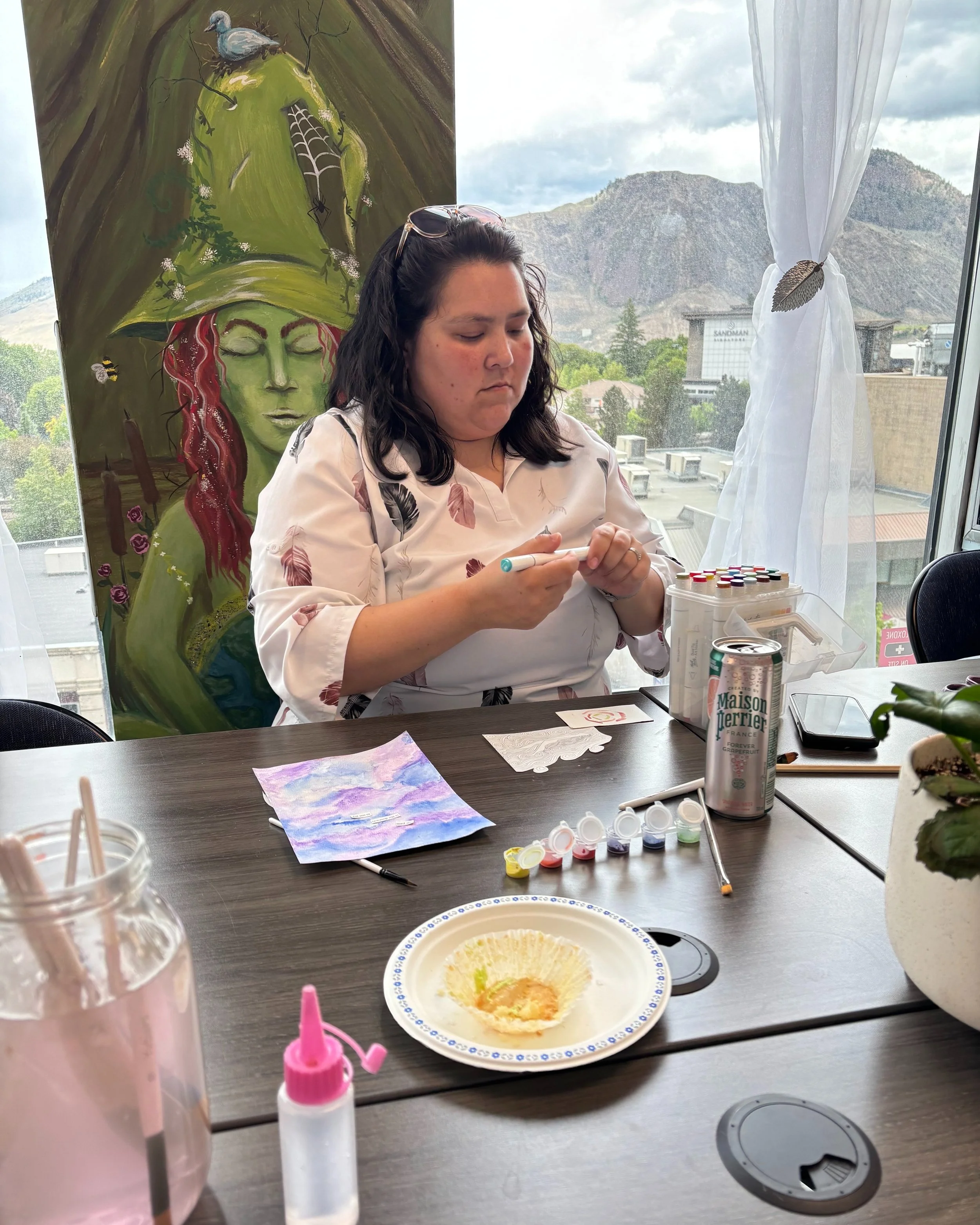 A woman with dark hair and light skin, sitting at a table, holding a small object in her hands. The table has art supplies, a water cup, a can of Maison Perrier, and a plate with some crumbs and remnants. Behind her is a window with sheer white curtains, and on the wall is a painting of a face with a green hat, a bird, and bees.