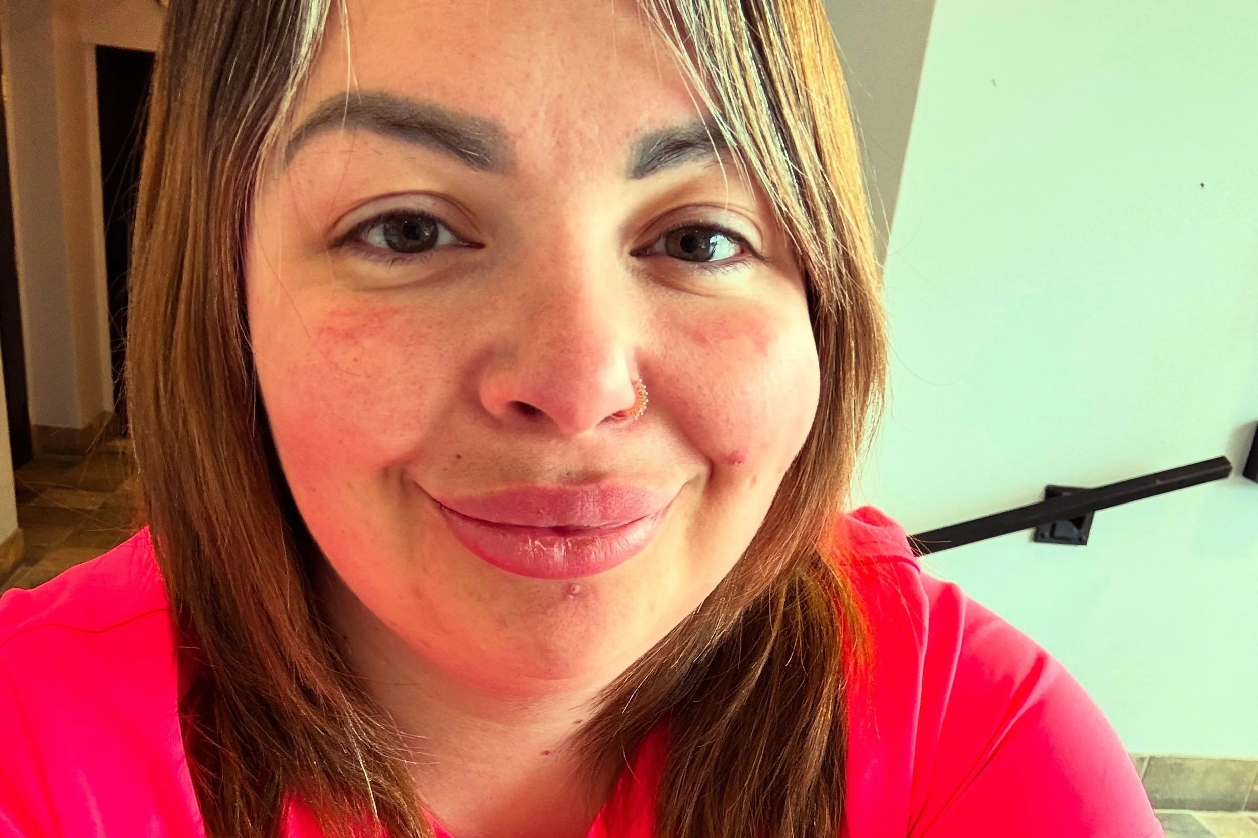 Close-up of a woman with shoulder-length brown hair, smiling, wearing a pink top, indoors with a white wall and a gray hallway in the background.