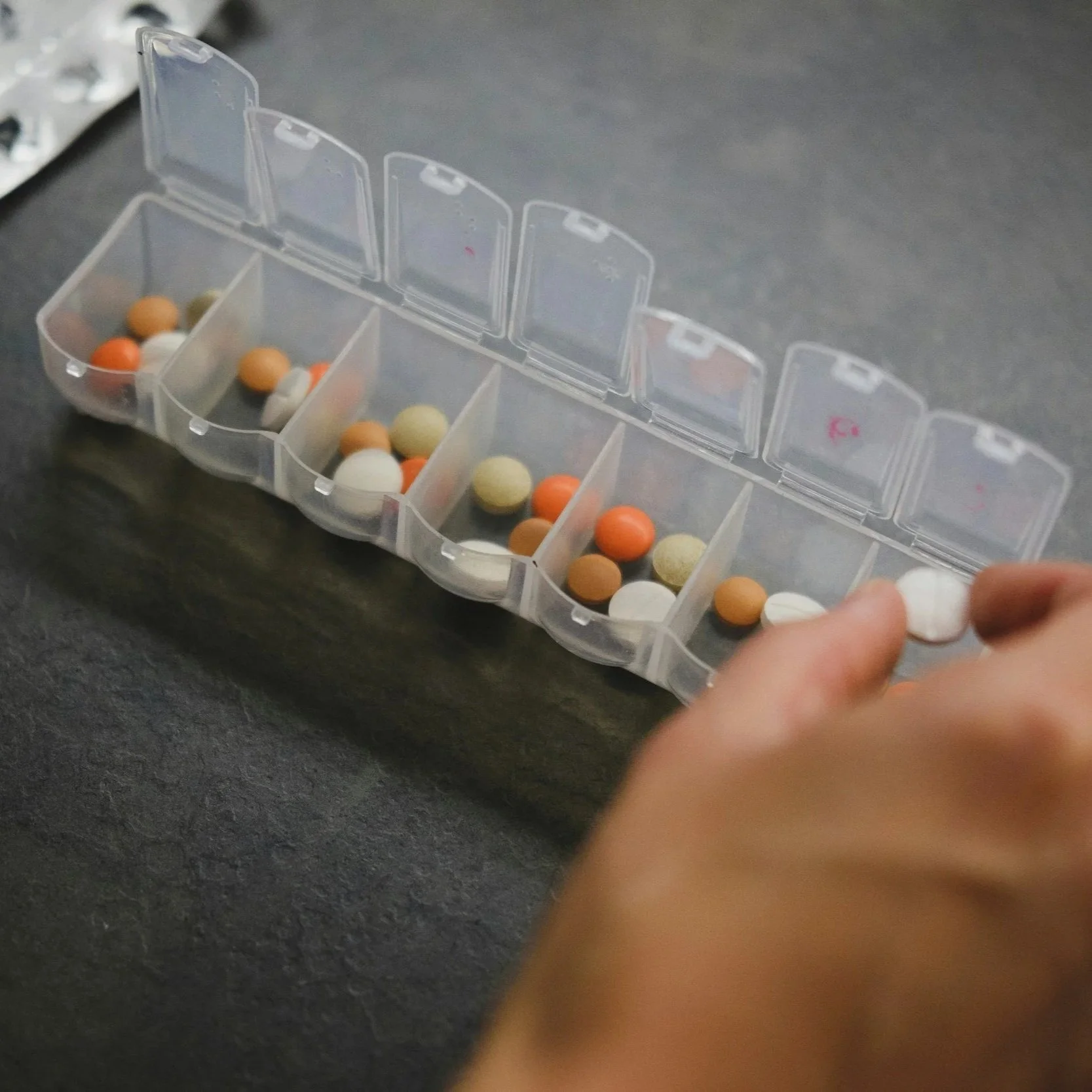 A pill organizer with multiple compartments containing various colored pills, with a person's hand reaching toward it on a dark surface.