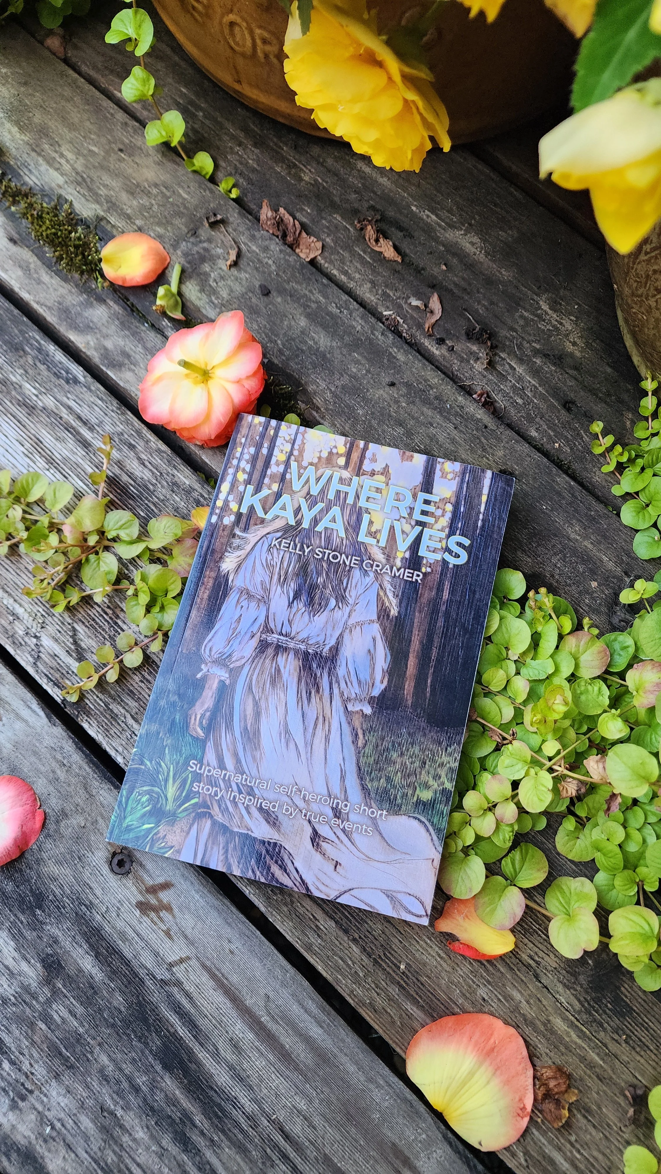 Book titled 'Where Kaya Lives' by Kelly Stone Cramer lying on grass with small white flowers.