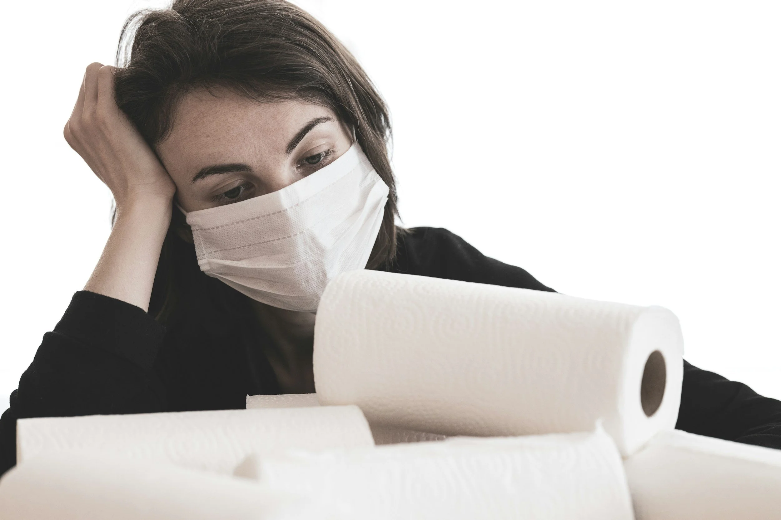 A woman with brown hair wearing a face mask, resting her head on her hand, and looking at several large rolls of white paper towels.