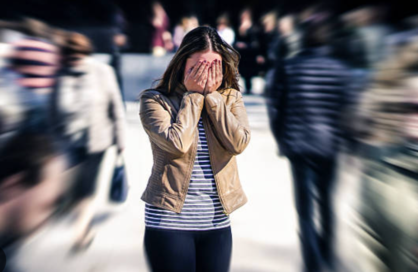 A woman with long dark hair covering her face with her hands in a busy outdoor area, surrounded by blurred people.