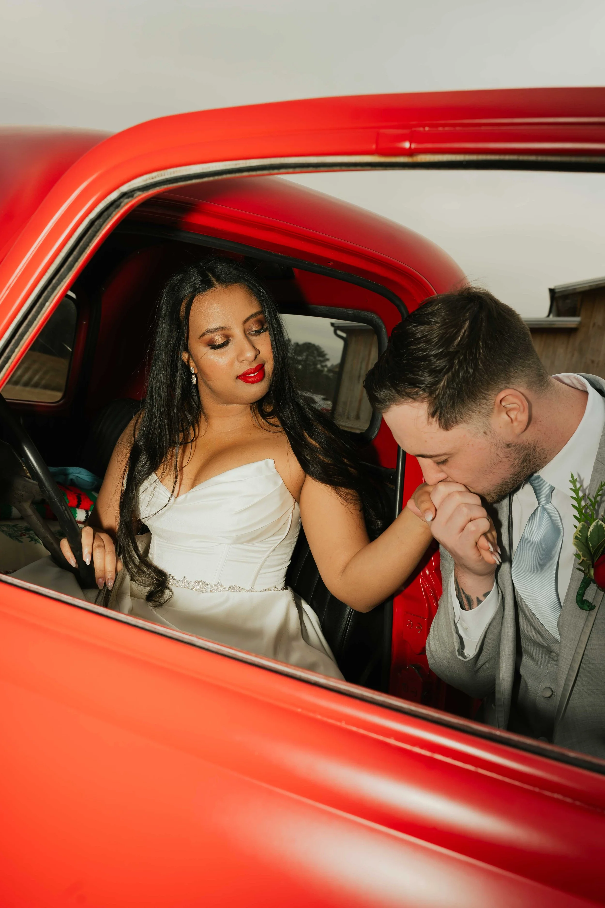A woman in a white wedding dress sitting in a red truck, holding the hand of a man in a gray suit inside the truck, with the man kissing her hand.